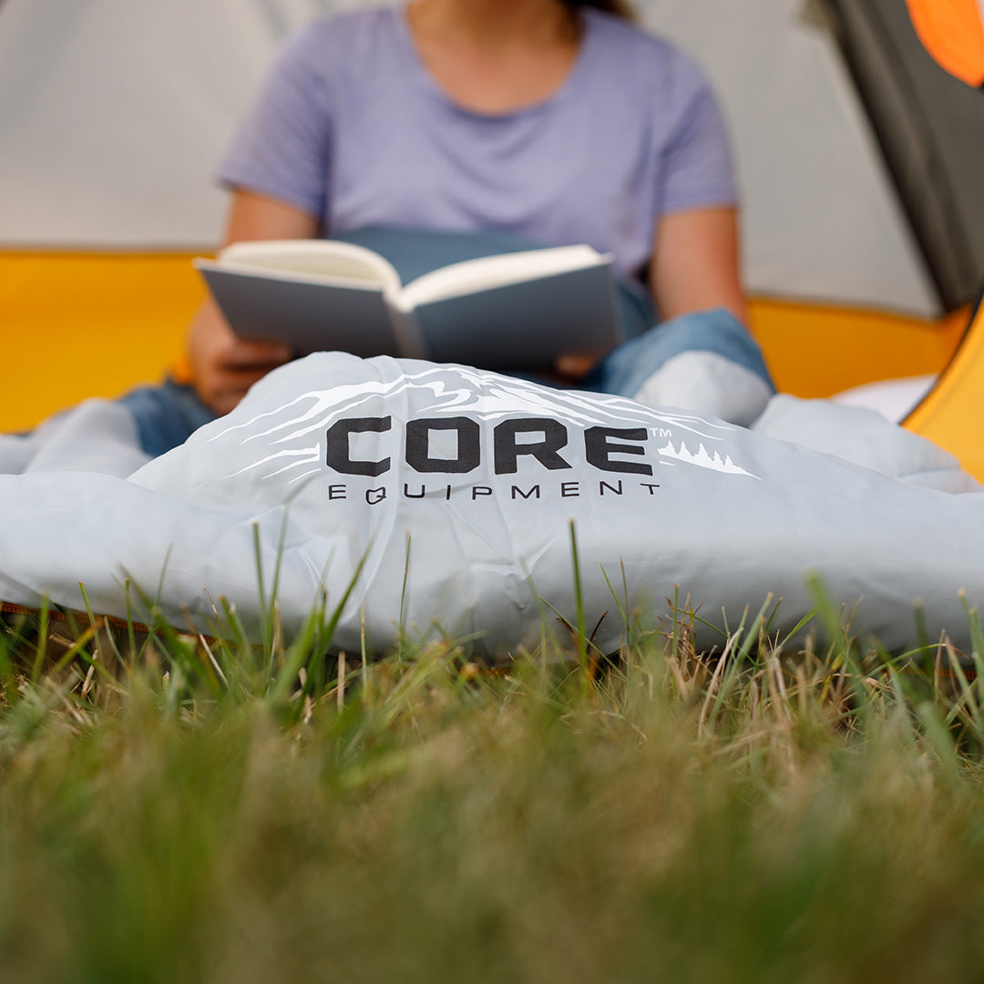 Camper reading inside a tent with the CORE 50 degree sleeping bag in gray, featuring soft lining and warm insulation.