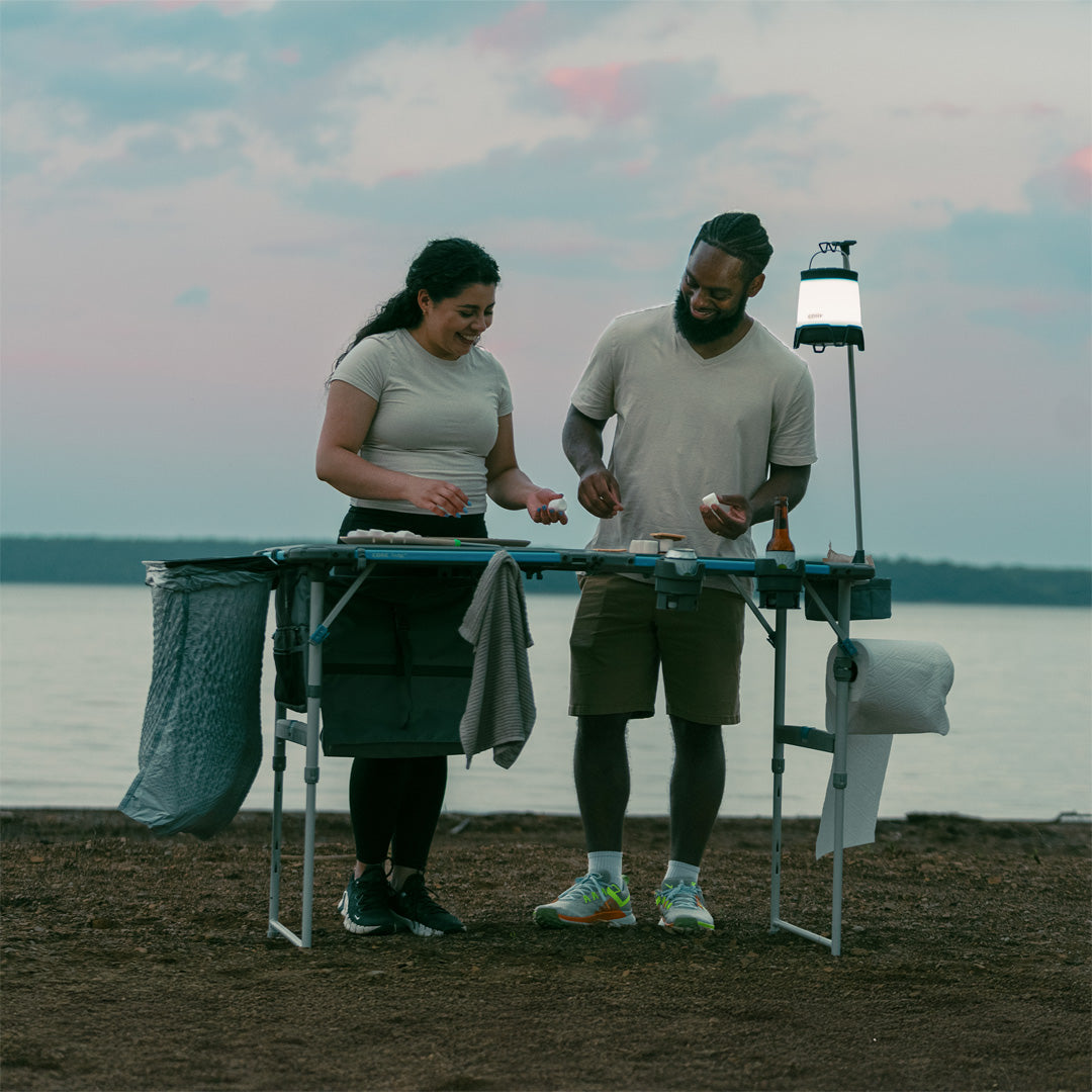 Campers cooking on the CORE 4 Foot FlexRail Folding Cook Table near a lake, featuring storage, lantern hook, and paper towel holder for organization.