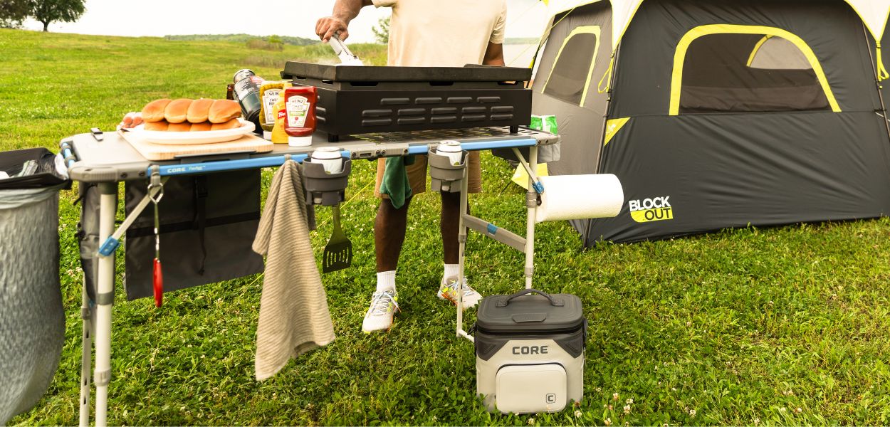 Camper cooking on the CORE 4 Foot FlexRail Folding Cook Table near a lake, featuring storage, lantern hook, and paper towel holder for organization.