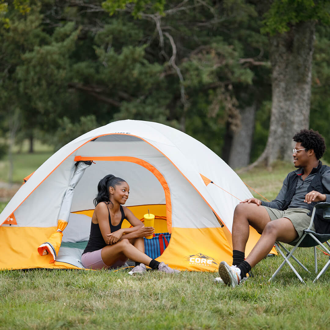 Campers relaxing beside the CORE 3 Person Dome Tent in gray and orange, designed for easy setup and comfortable outdoor shelter.