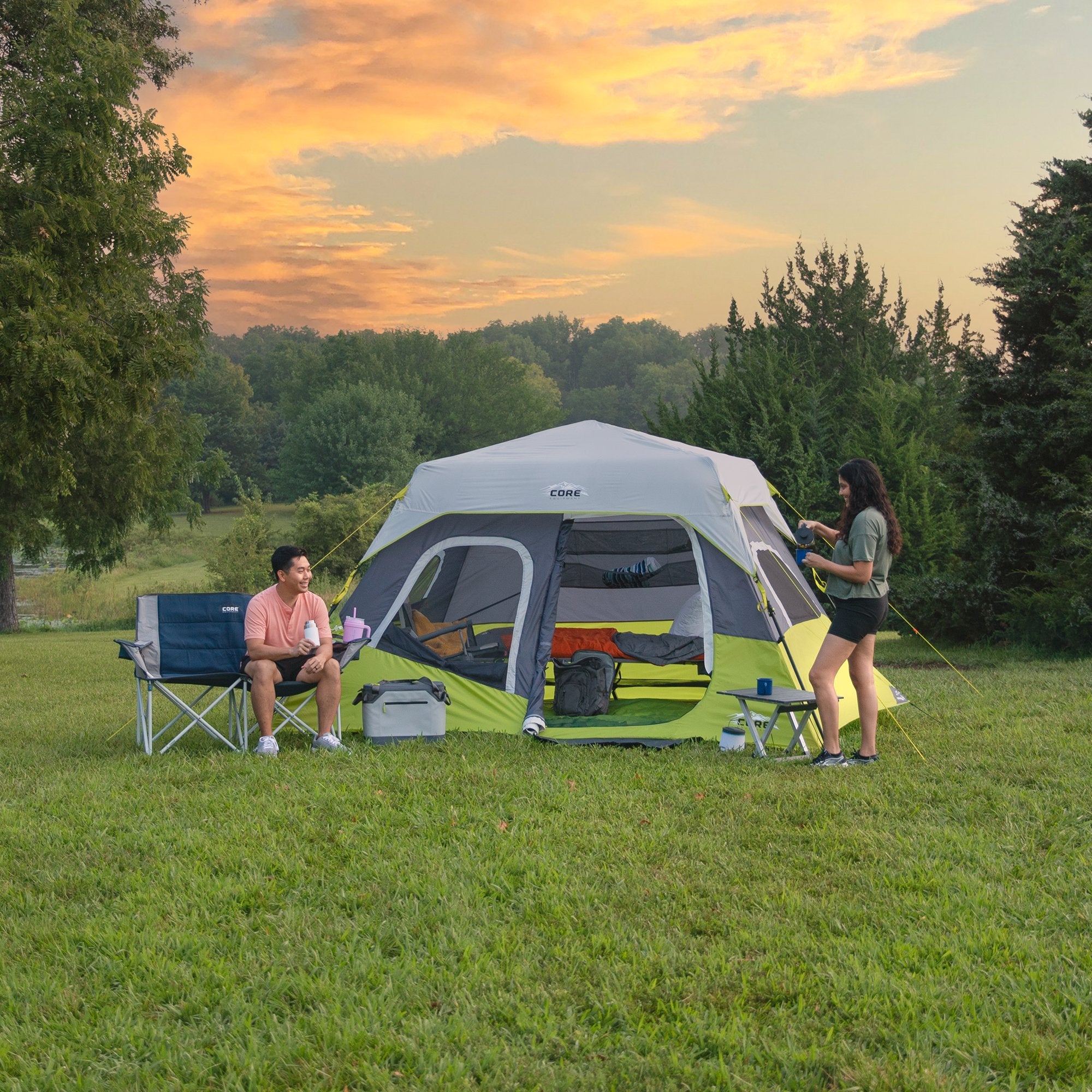 Campers relaxing beside the CORE 6 Person Instant Cabin Tent in gray and green, featuring quick setup and roomy interior space.