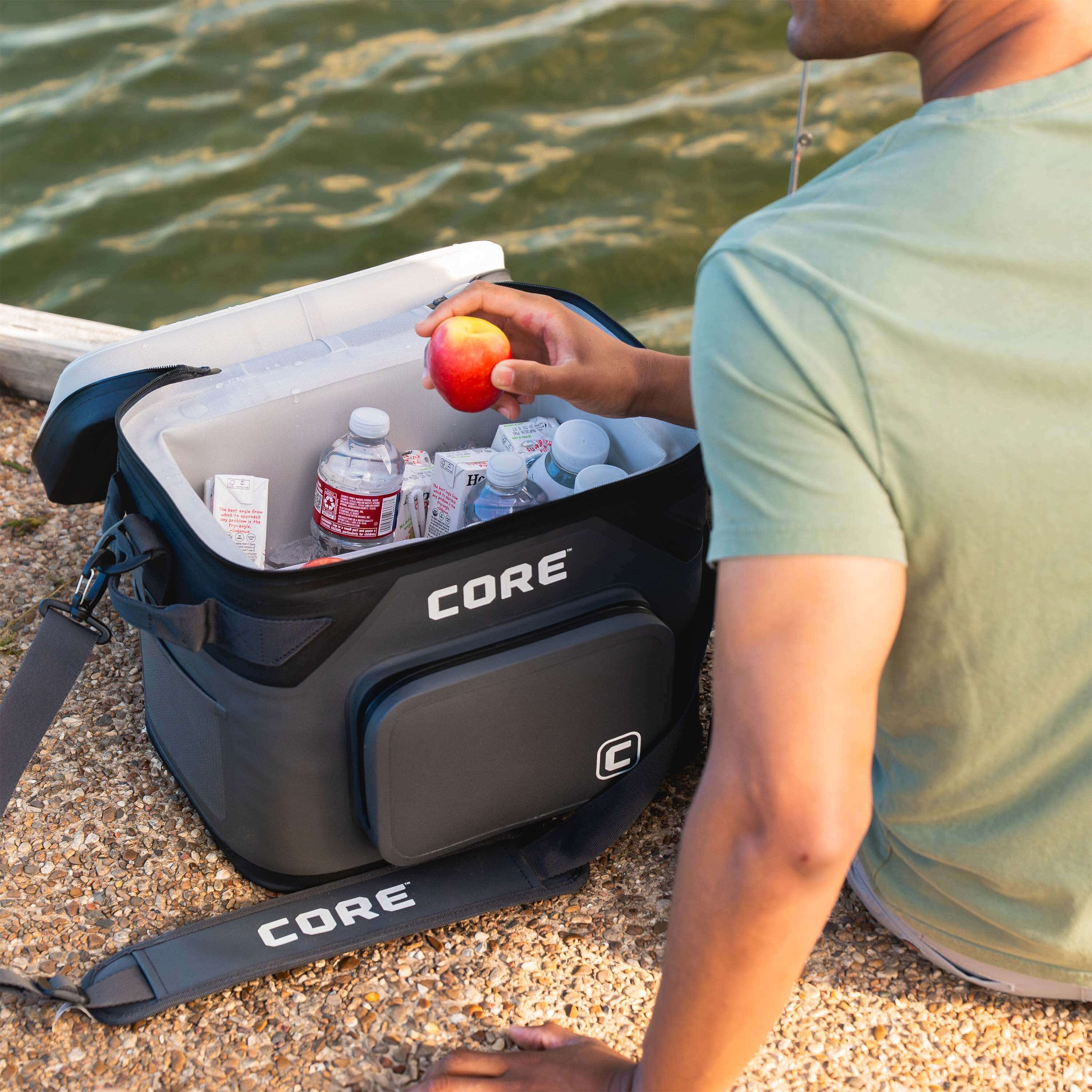 Person sitting by the water with a CORE Climakeep 18 can soft cooler filled with drinks and snacks, featuring shoulder strap and front pocket.