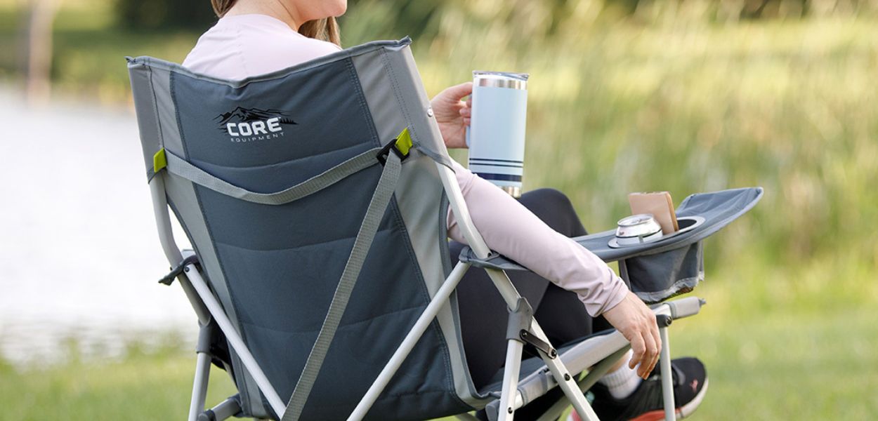 Camper relaxing by the water in the CORE Tension Chair in gray, featuring built-in cup holder and sturdy folding frame design.