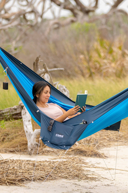 A person is reading a book in a blue double premium hammock outdoors.