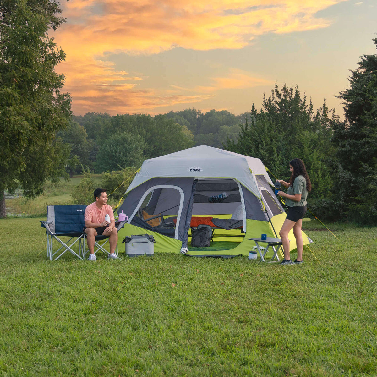 Two people camping outdoors with a CORE 6 person instant cabin tent in a grassy area during sunset.