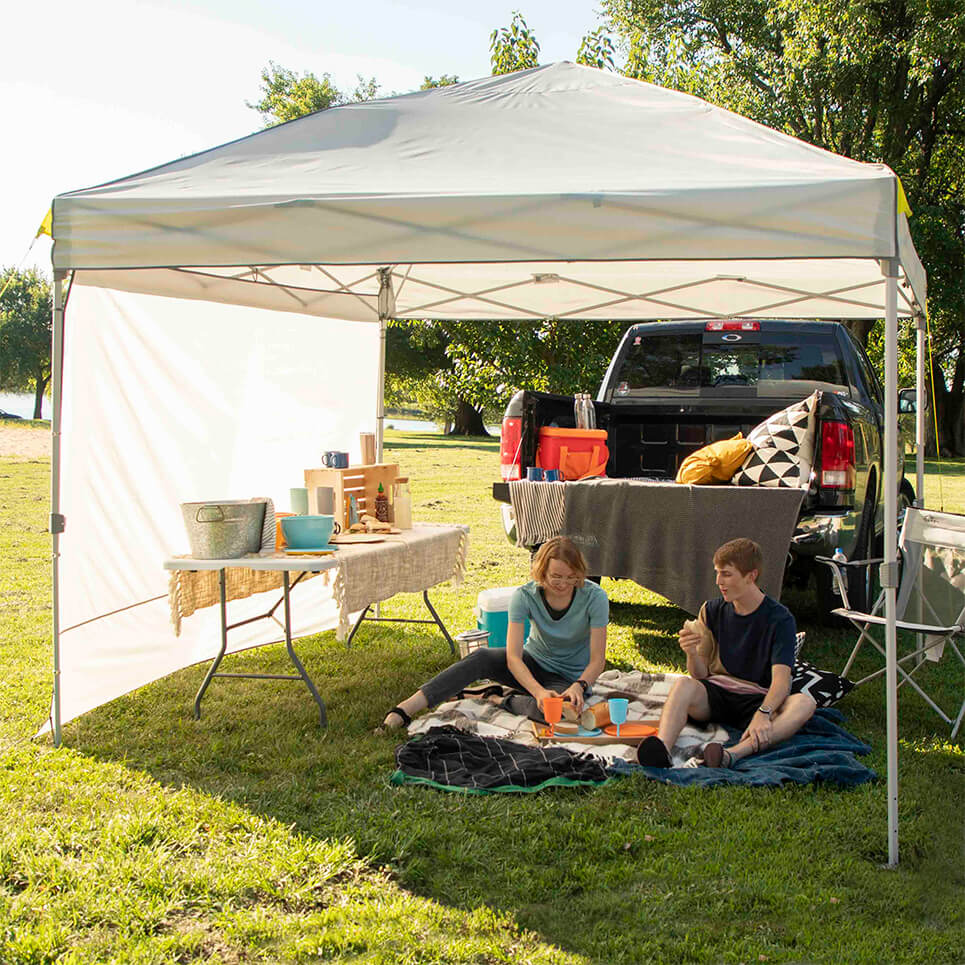 Two people sit under a gray canopy tent in a park setting, with a truck in the background.