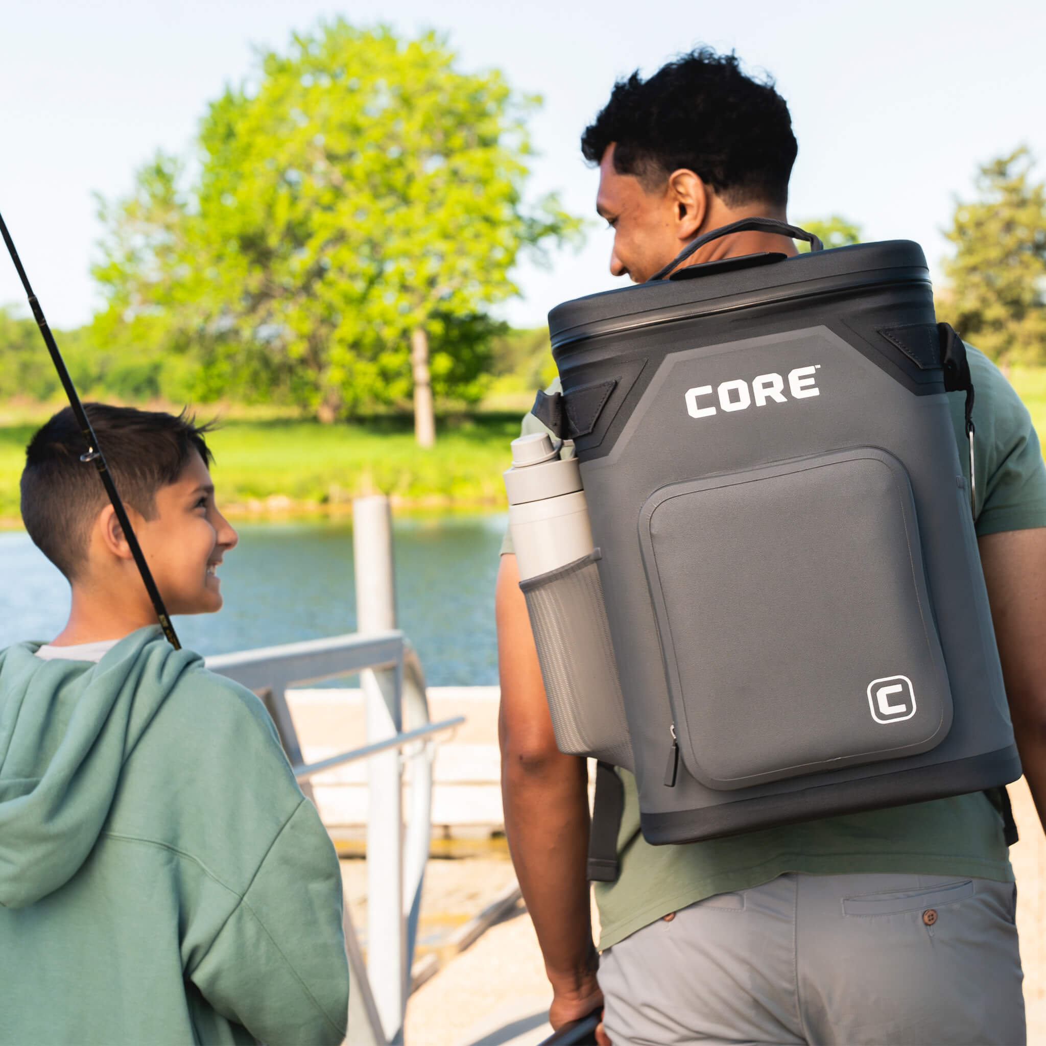 Man carrying CORE Climakeep 24 can charcoal backpack cooler with shoulder strap and side bottle holder while fishing with child.