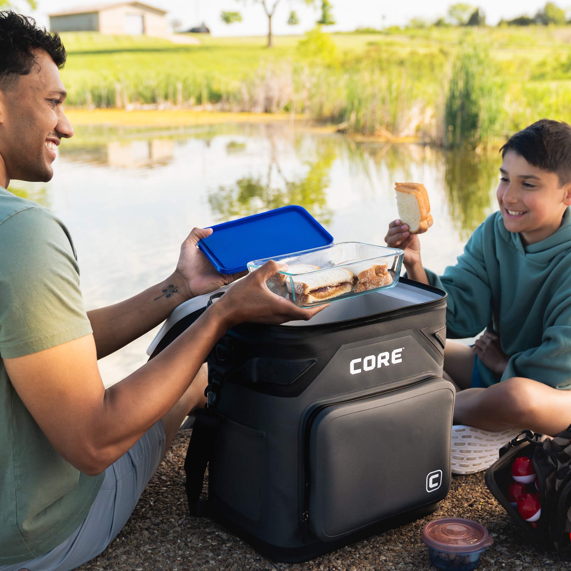 Two campers using a CORE Climakeep 36 can charcoal cooler by the water while eating outdoors.