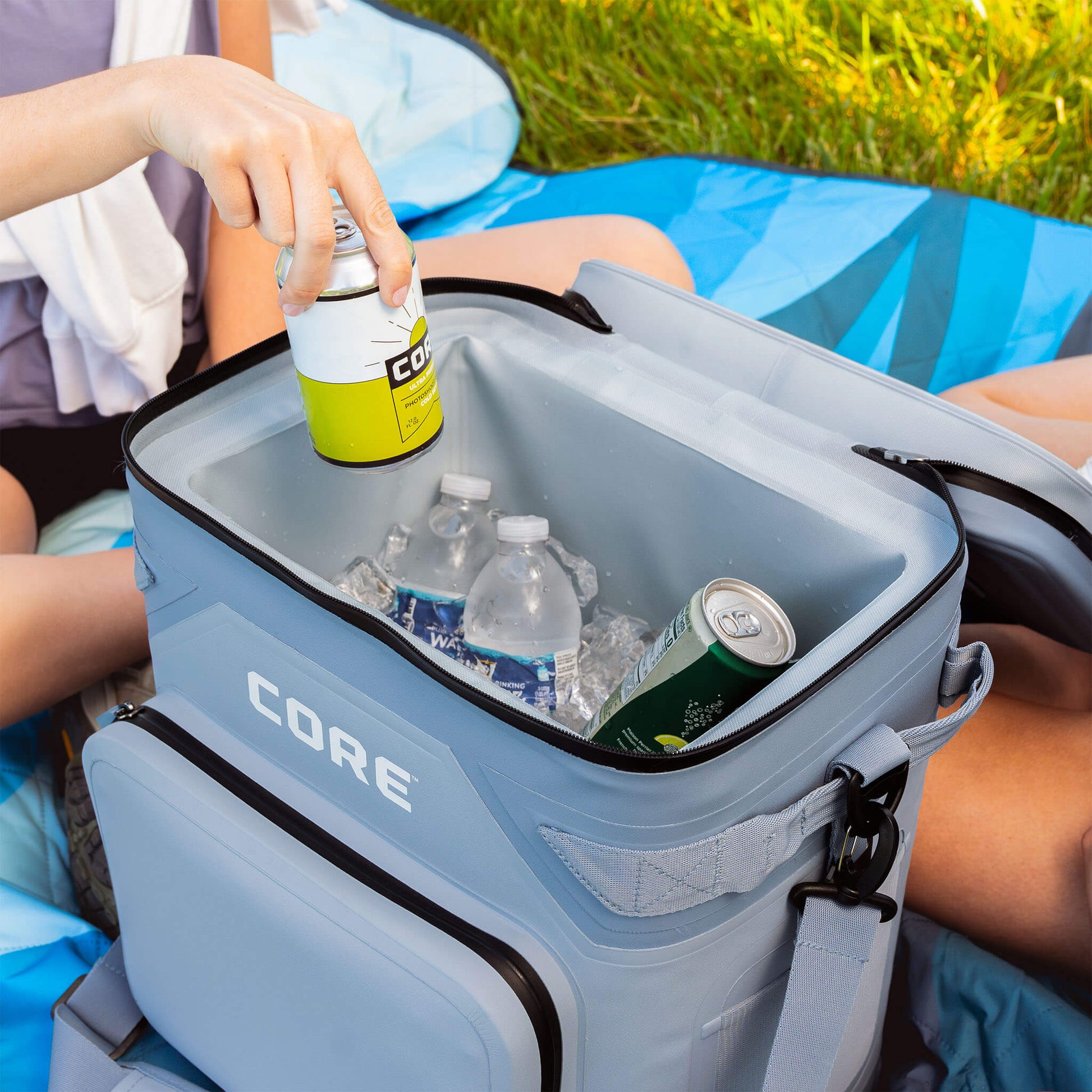 Person placing a can into an open CORE 36 Climakeep steel blue cooler, filled with ice, cans, and water bottles.