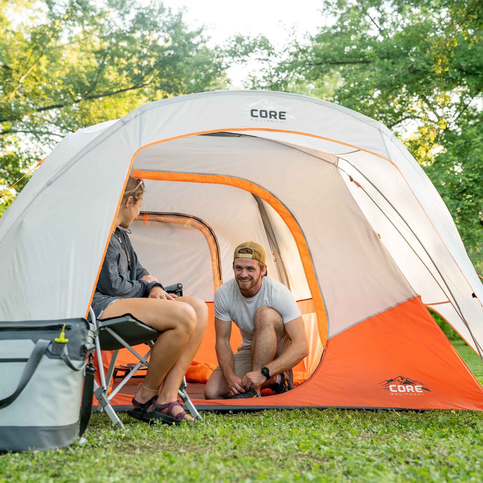 Two people sitting inside a CORE 6 person dome tent with a vestibule in a grassy area with trees.