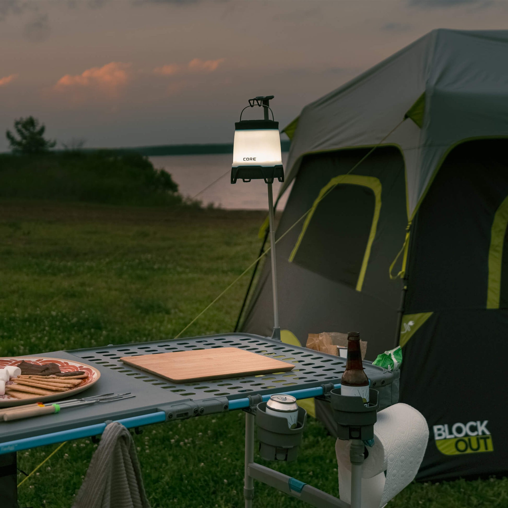 Lantern hanging from CORE lantern holder beside tent and FlexRail table at dusk, providing convenient hands-free outdoor camp lighting.