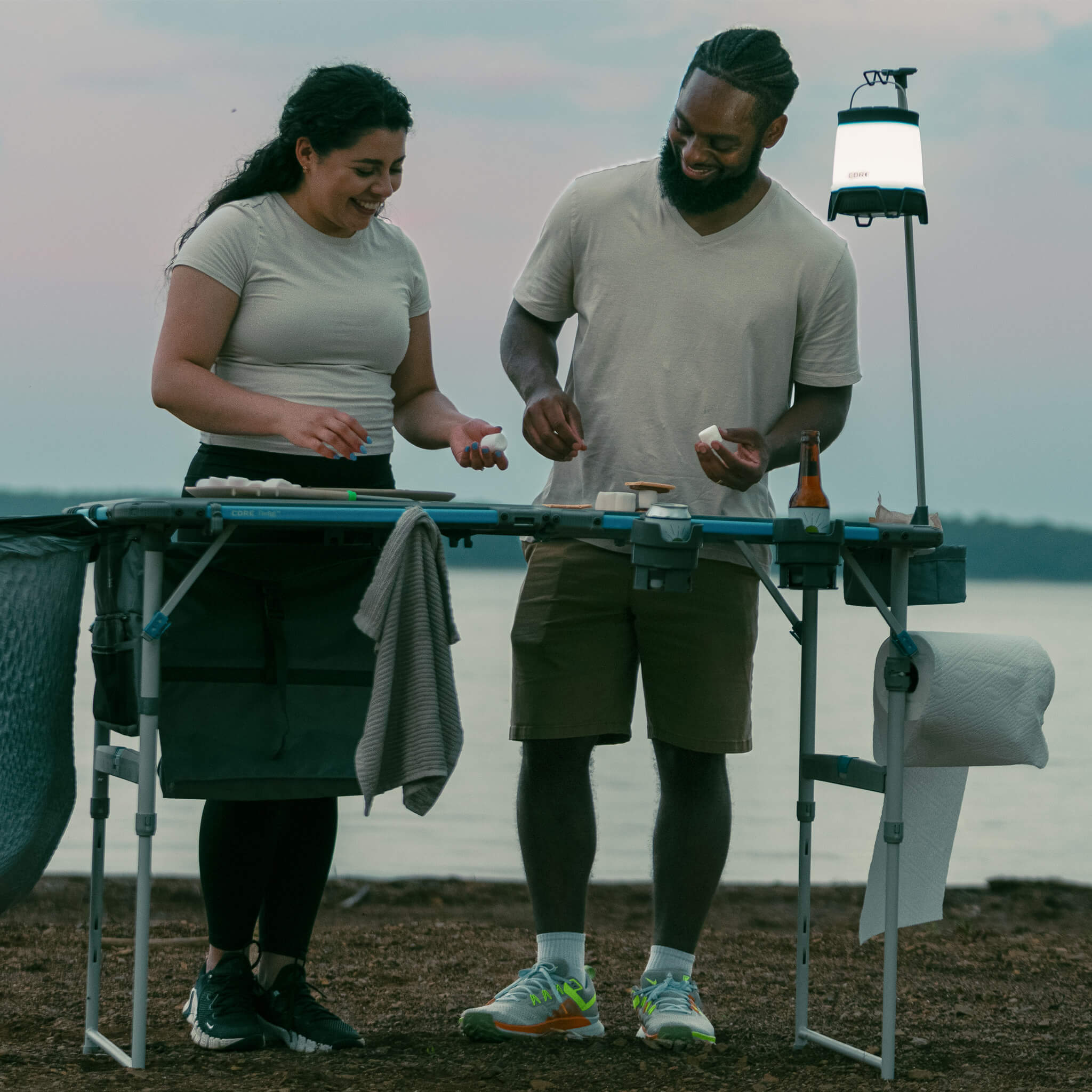 Two campers cooking beside table with CORE lantern holder providing hands-free lighting, ideal for evening outdoor camp activities.