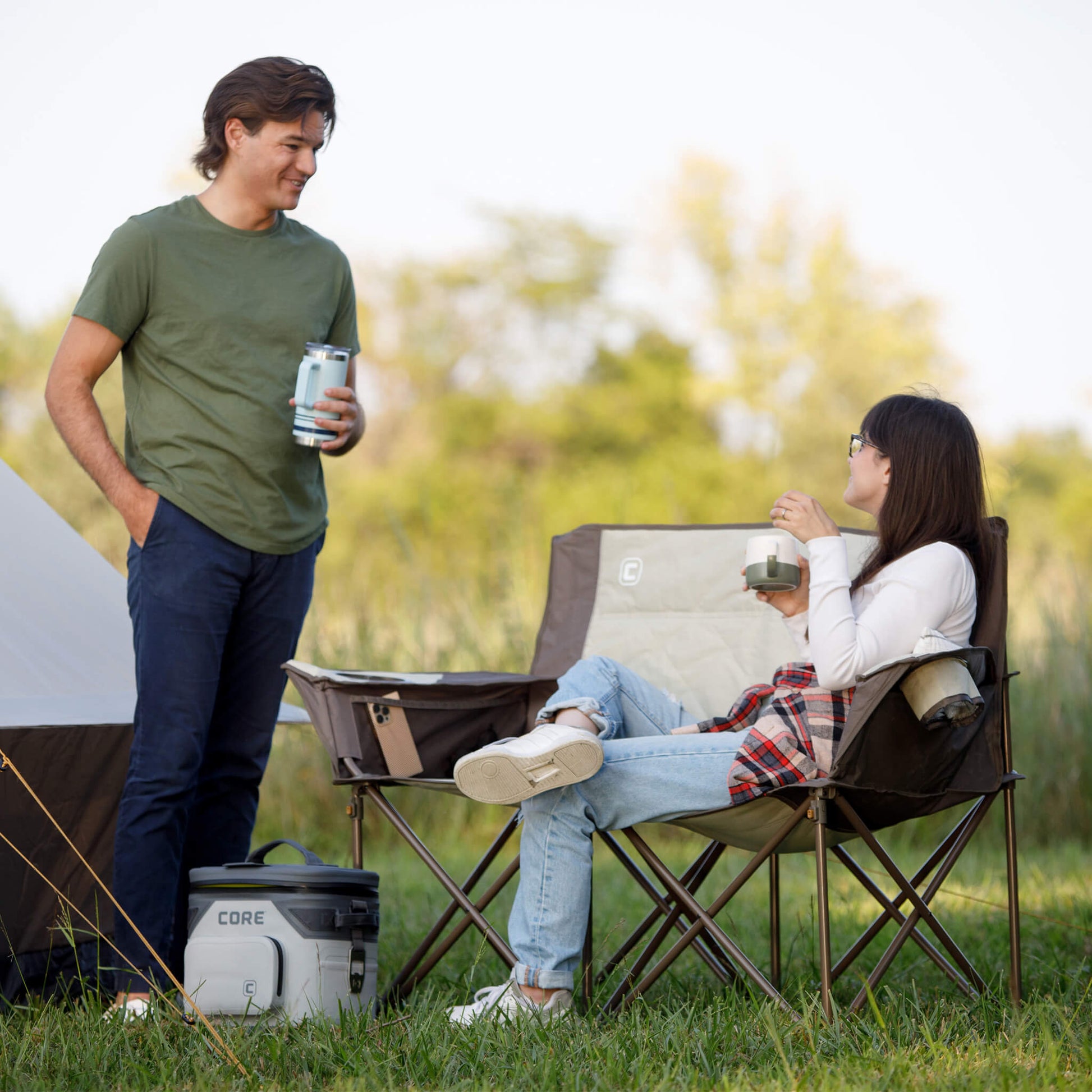 Two campers relaxing by a tent using the CORE loveseat camp chair in tan and brown.