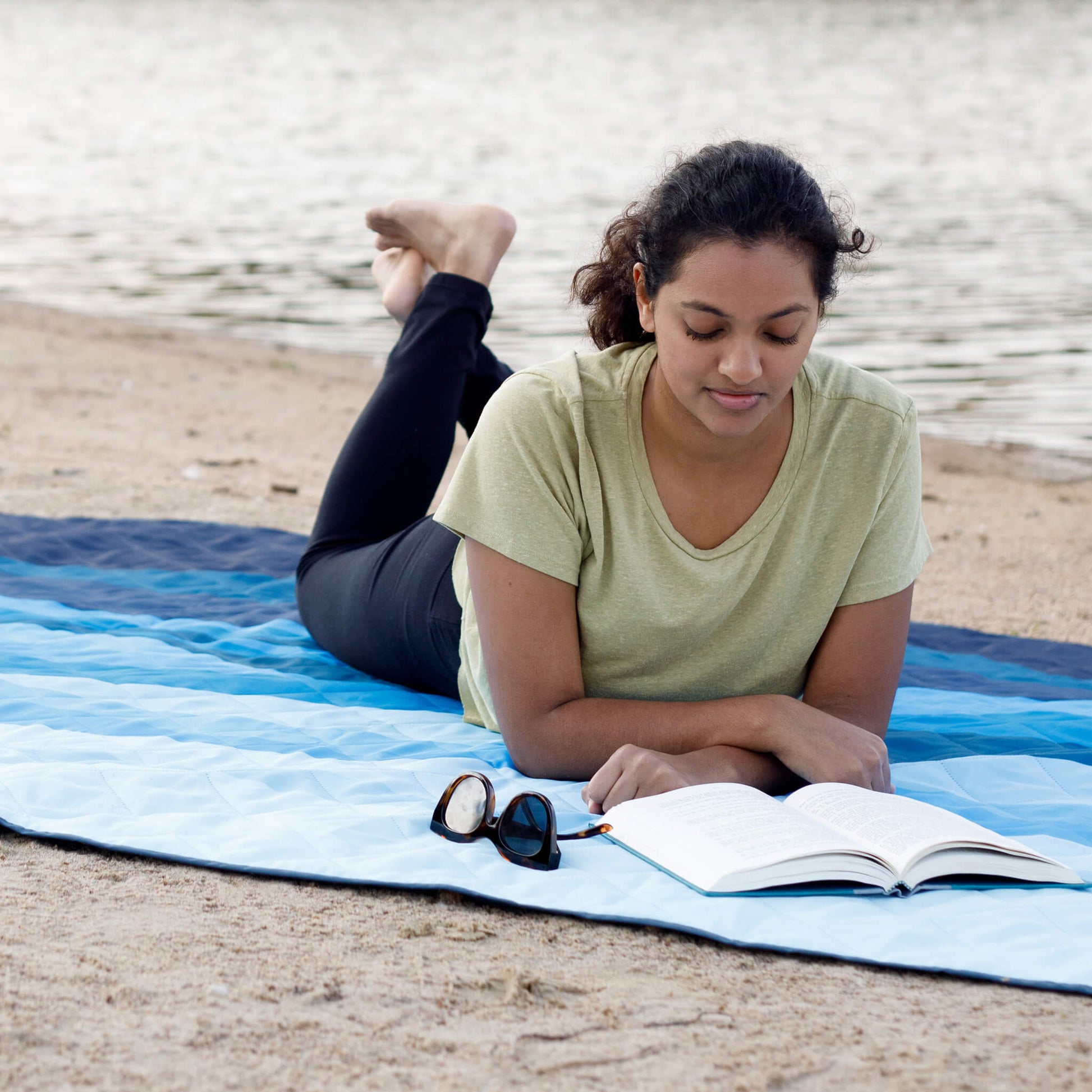 A women lying on a CORE outdoor blanket in blue pattern while reading outside.