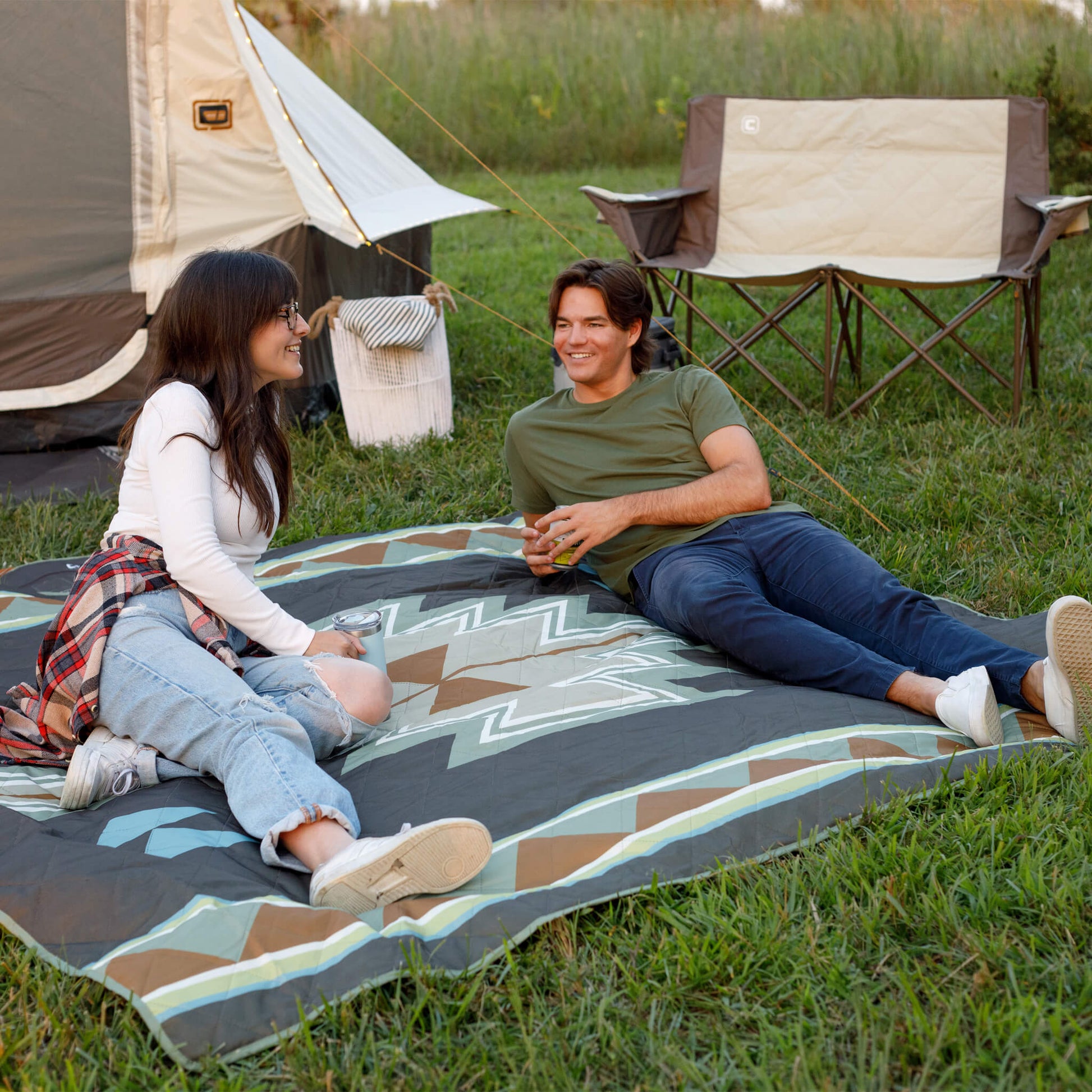 Two people sitting on a brown southwestern desert mosaic patterned camping blanket in a grassy area with a tent and chair in the background.