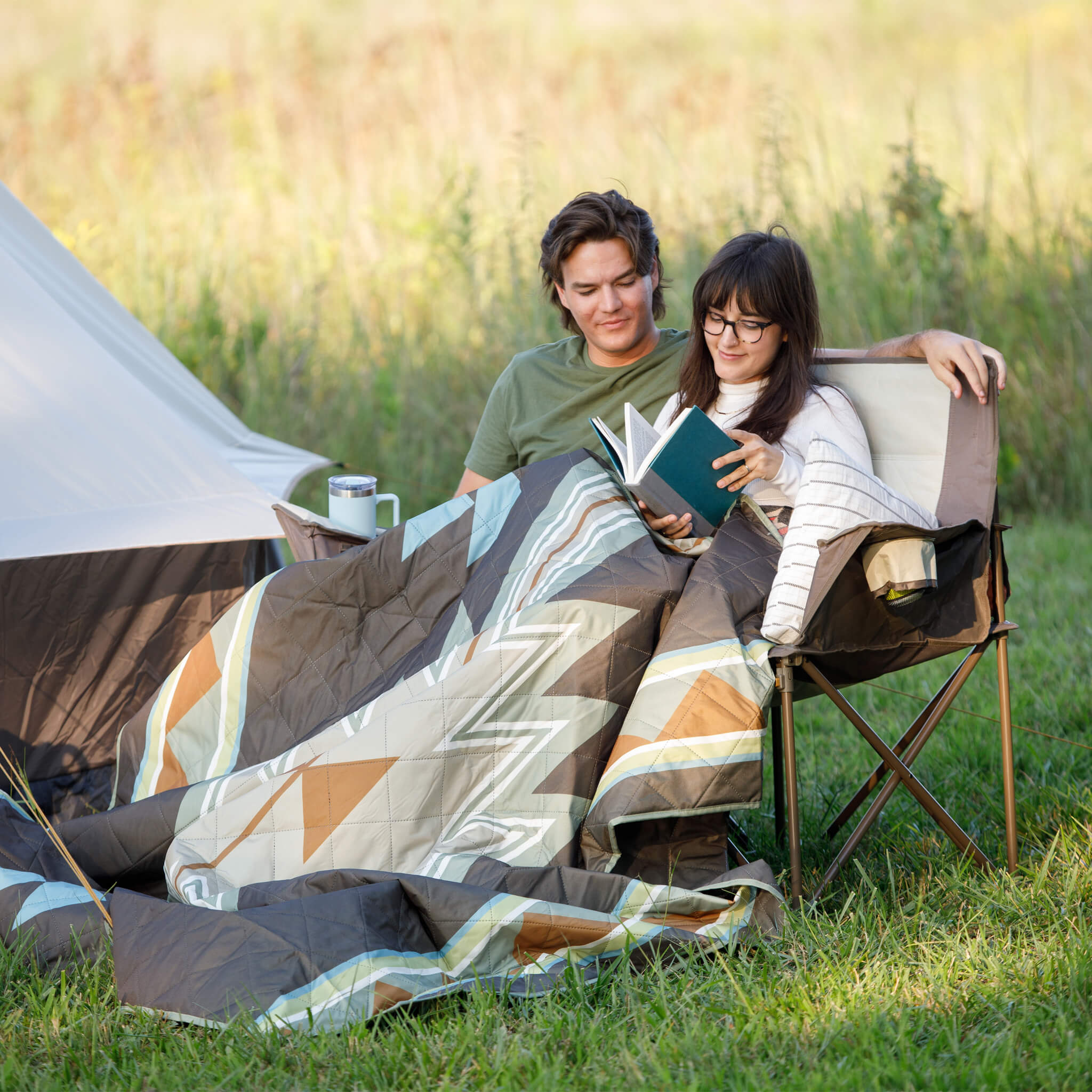 Two people are sitting under a southwestern desert mosaic patterned blanket in a grassy field with a tent in the background.