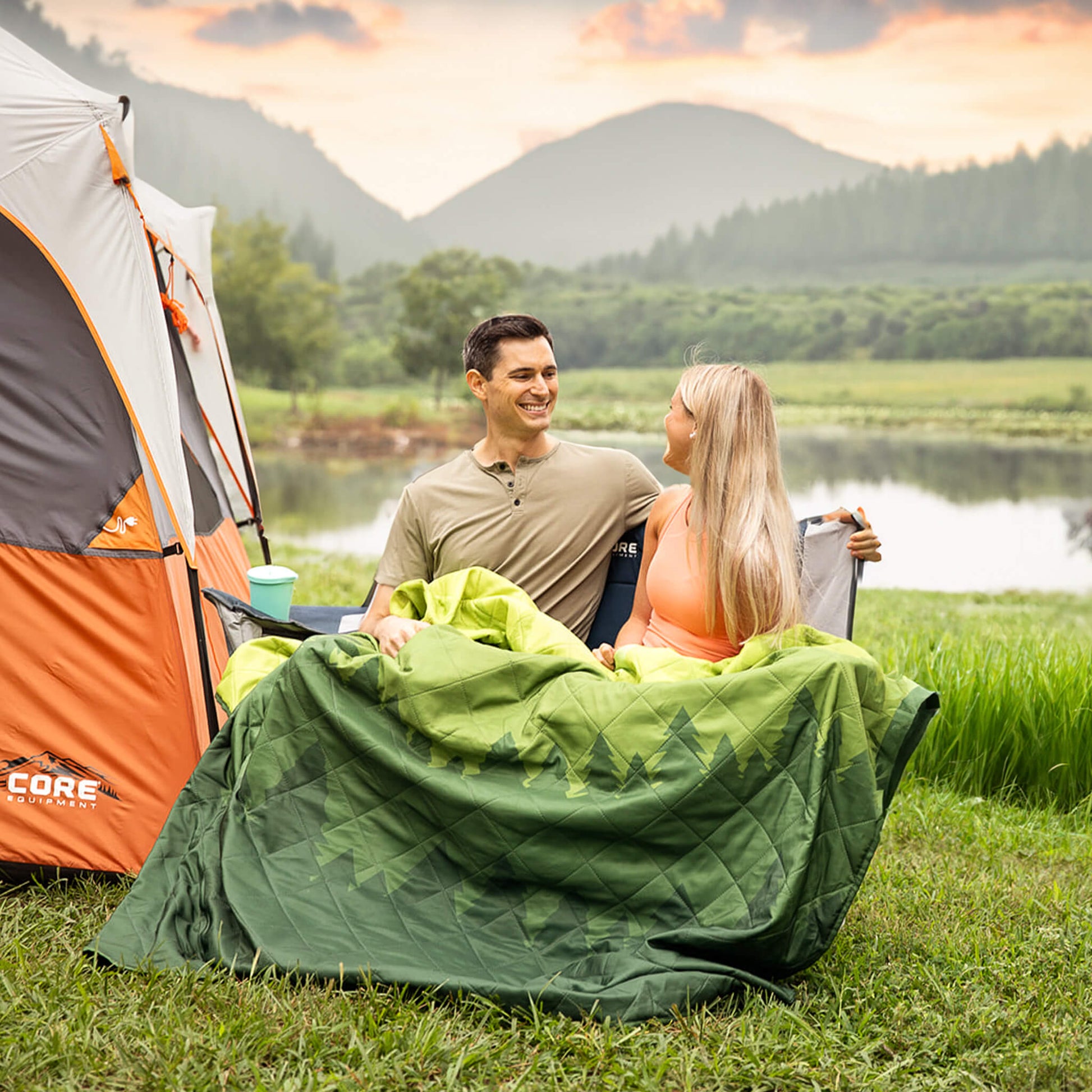 A couple sitting at a campsite in a loveseat chair with the CORE green weighted outdoor blanket in front of a tent.