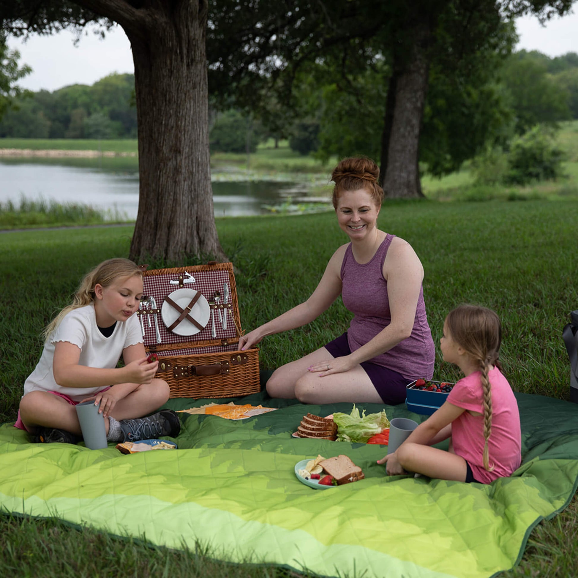 Family sitting on a CORE green weighted outdoor blanket having a picnic in the park.