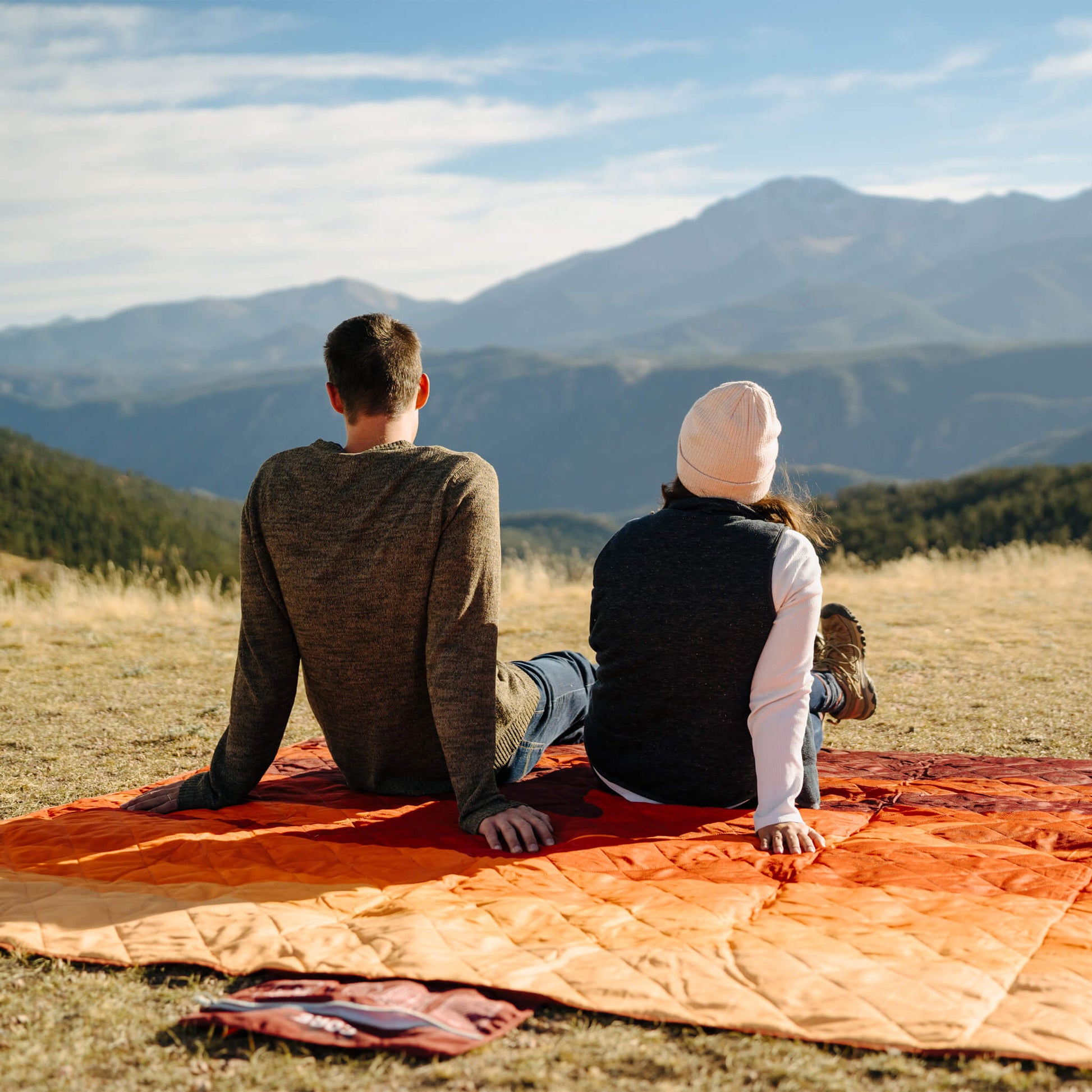 A couple sitting on a CORE outdoor blanket in orange pattern with mountain view.