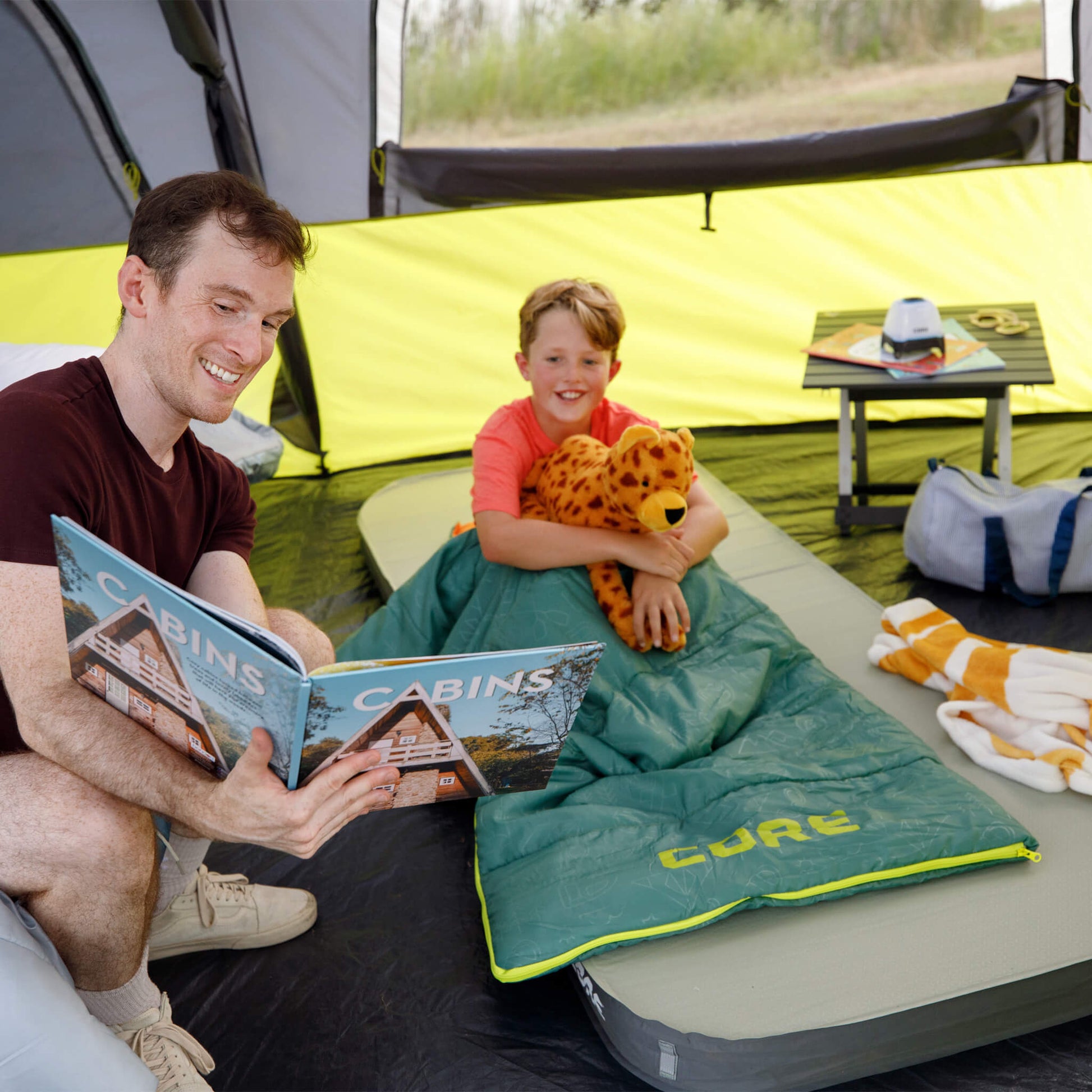 A father reading with a child using a green CORE Youth 50 Degree sleeping bag inside a CORE instant tent.