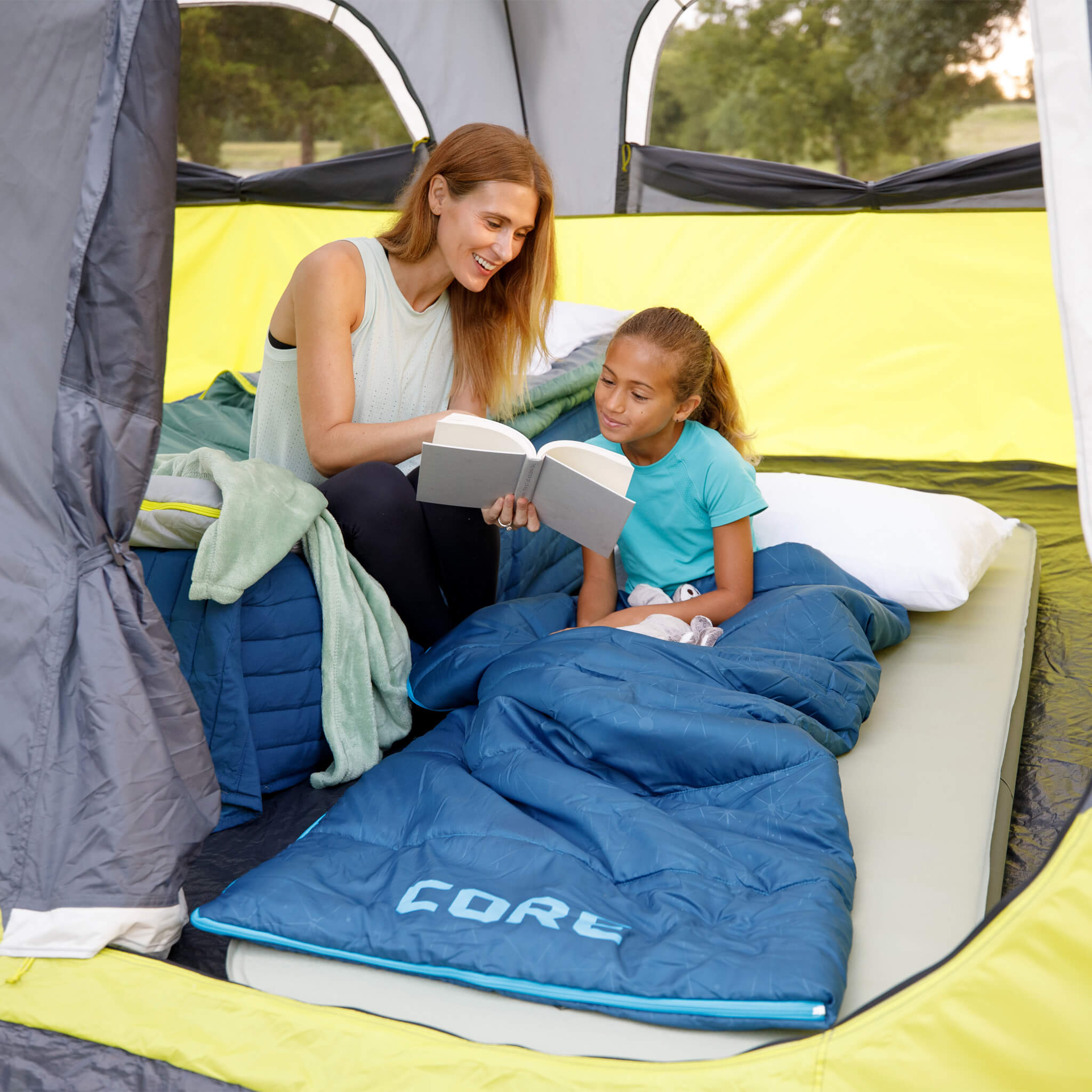 A mother reading to a daughter in a CORE instant tent using a CORE Youth 50 Degree blue sleeping bag.