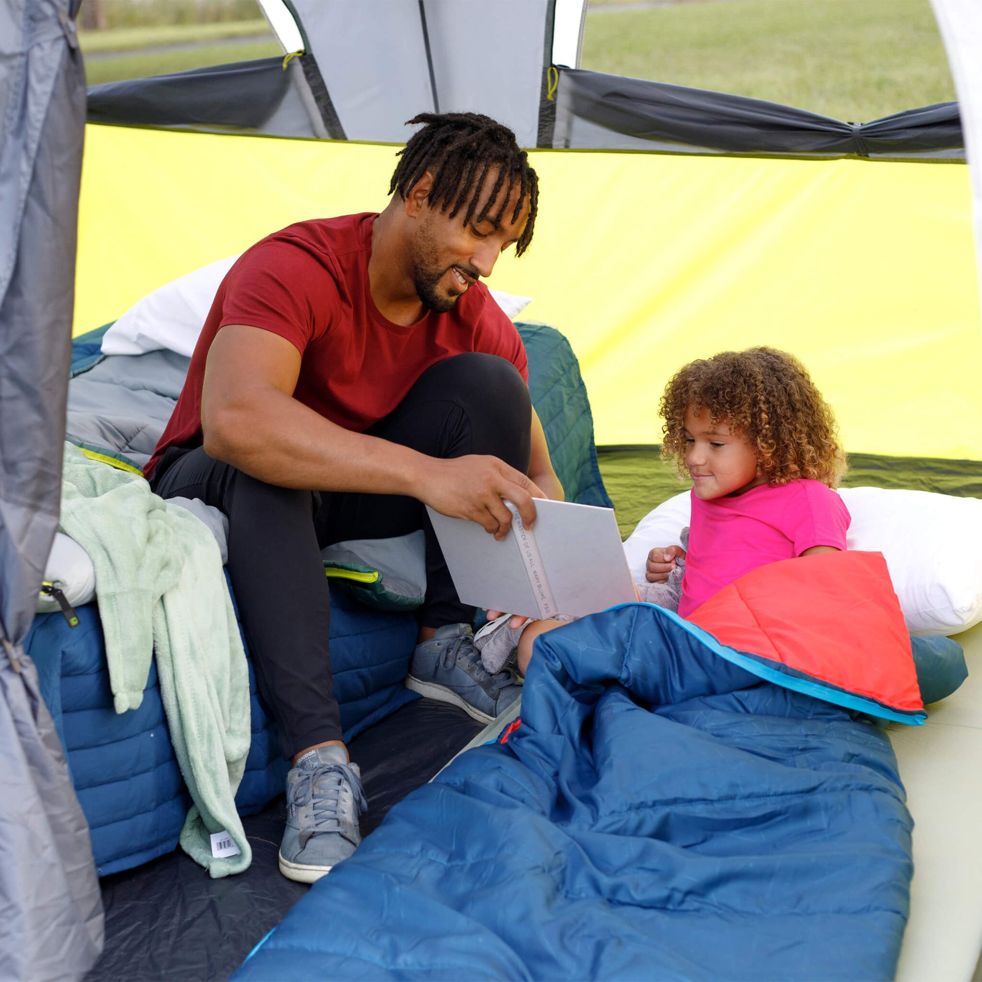 A father reading with a child using a blue CORE Youth 50 Degree sleeping bag inside a CORE instant tent.