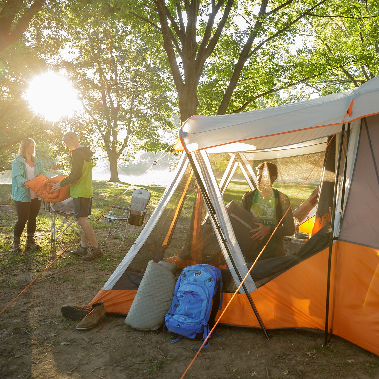A group of people is setting up a campsite in a forested area. They are standing by an orange and grey CORE 11 Person Cabin Tent with Screen Room. The sun is shining through the trees, and various camping equipment, including a blue backpack, is scattered around. One person is holding a sleeping bag.