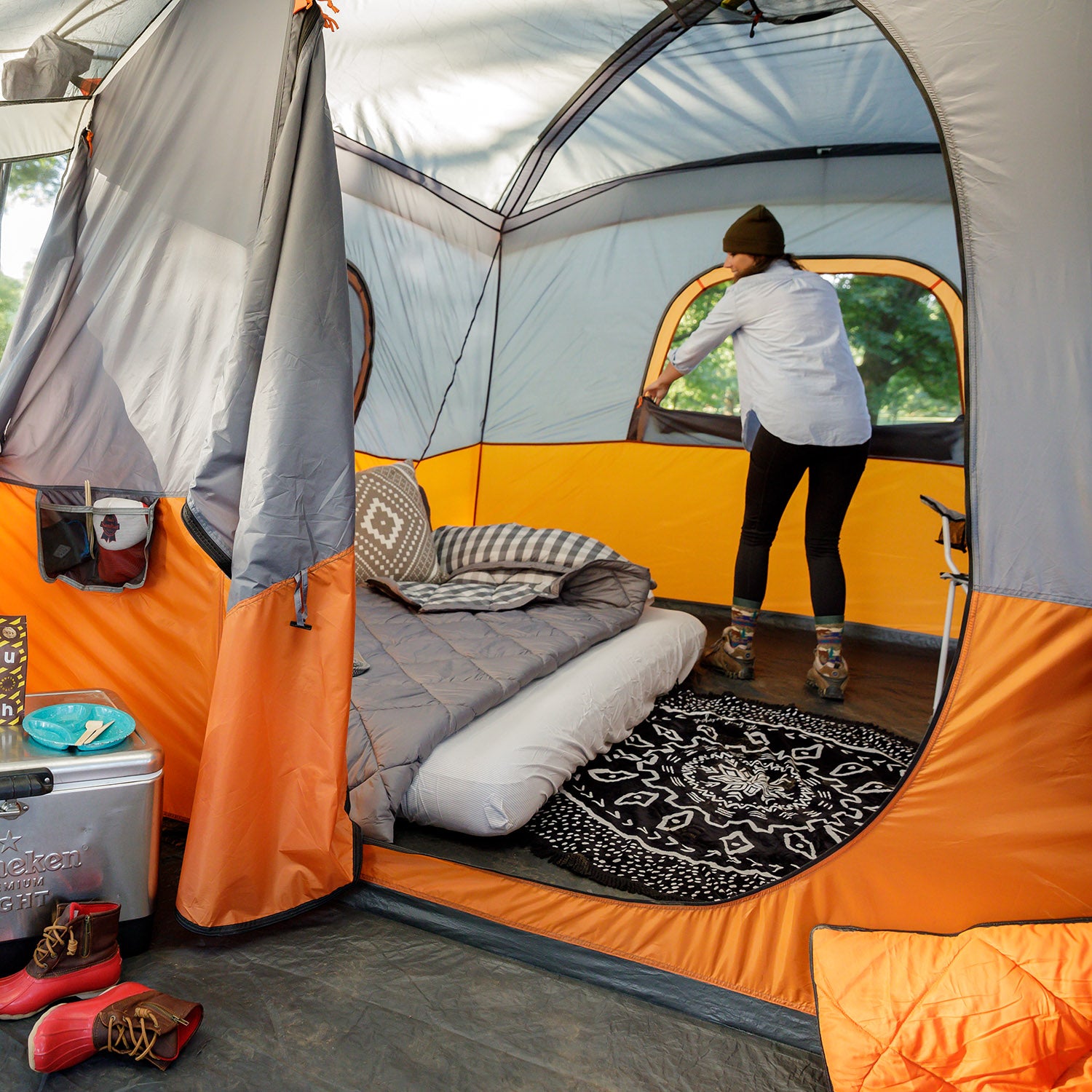 A person wearing a beanie and jacket is seen adjusting the window flap of a brightly colored CORE 11 Person Cabin Tent with Screen Room. Inside the tent, there's an inflatable mattress with pillows and a blanket, along with a storage box, hiking boots, and a patterned rug on the floor.