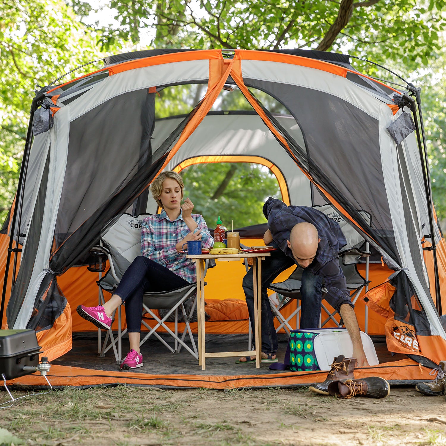 Two people are sitting in a large, open-front CORE 11 Person Cabin Tent with Screen Room surrounded by trees. One person is seated at a small table, wearing a plaid shirt and pink shoes, while the other person is bending down, wearing a blue shirt and reaching for an item on the ground.
