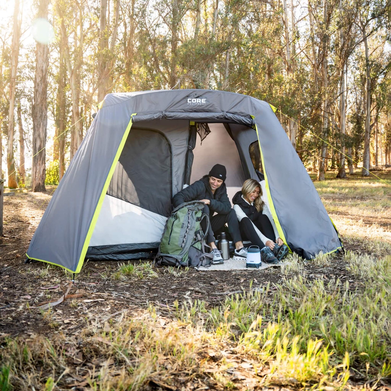 A man and woman sitting on the ground near the entrance of their CORE full rainfly camping tent. On the left, a man is digging through his hiking backpack. On the right, the woman is tying her shoes.
