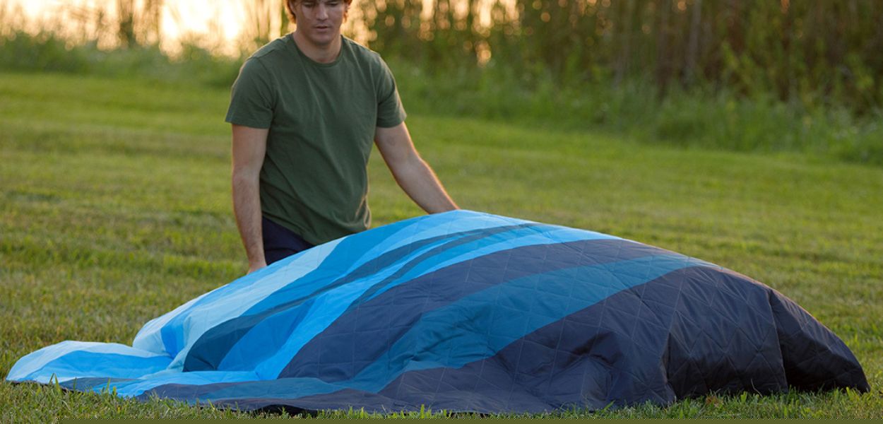 A camper laying out a CORE Staydown weighted outdoor blanket in blue on a grassy field at sunset.