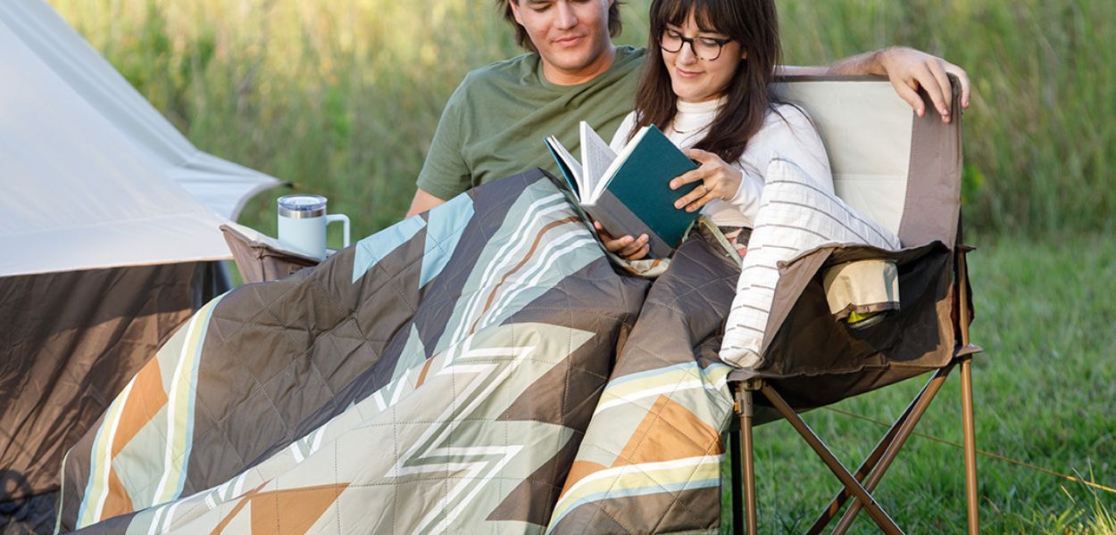 Couple relaxing outdoors under a patterned weighted camping blanket near a CORE tent, reading together on a camping chair.