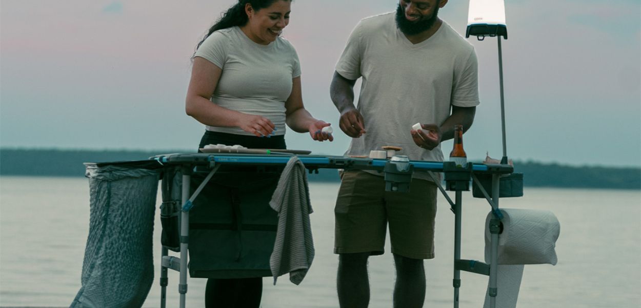 Campers cooking on the CORE 4 Foot FlexRail Folding Cook Table near a lake, featuring storage, lantern hook, and paper towel holder for organization.