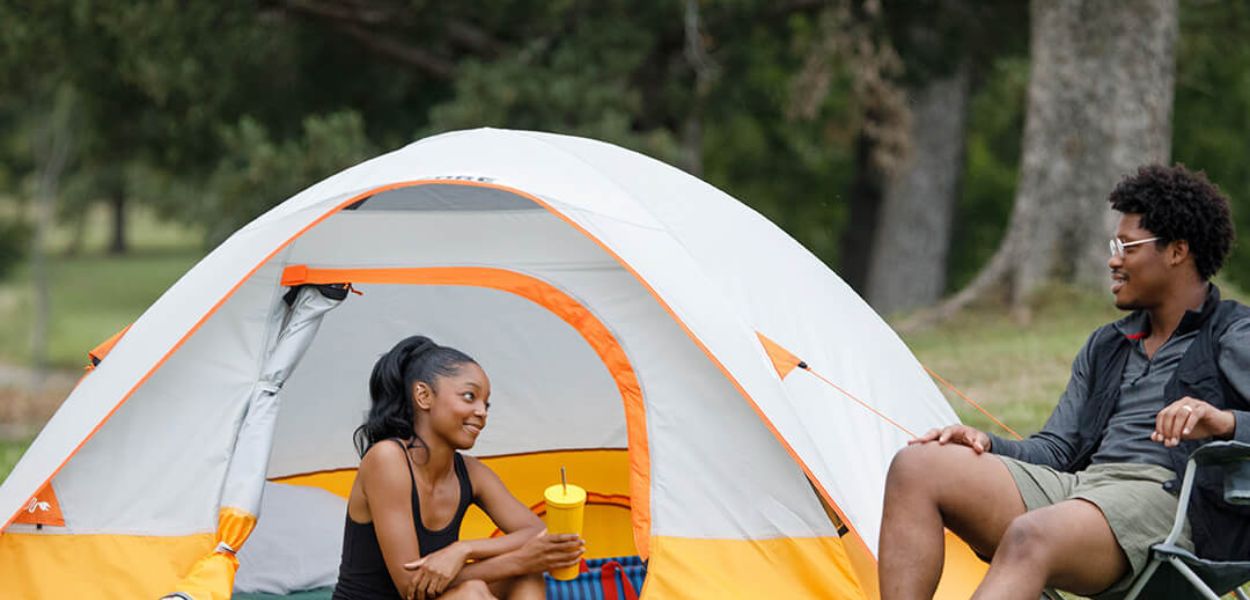 Campers relaxing beside the CORE 3 Person Dome Tent in gray and orange, designed for easy setup and comfortable outdoor shelter.