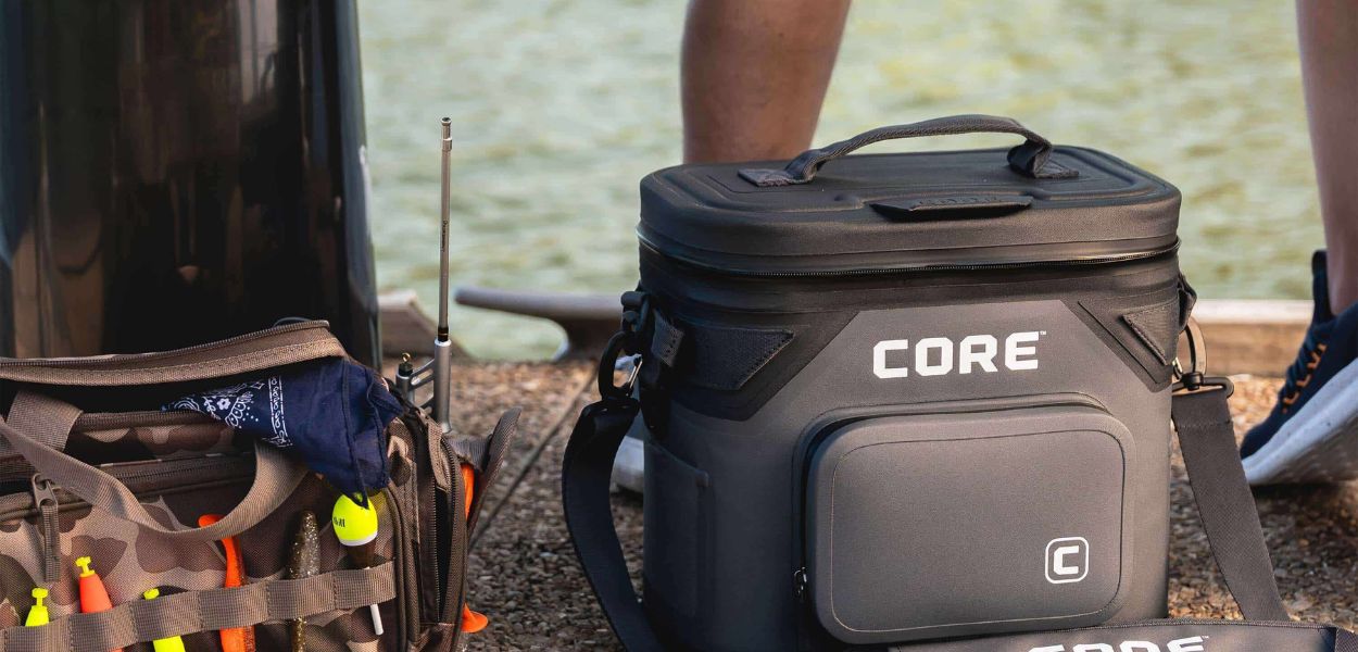 Person sitting on a dock with fishing gear beside a CORE Climakeep soft cooler in charcoal, shown next to a tackle box and fishing net near the water.