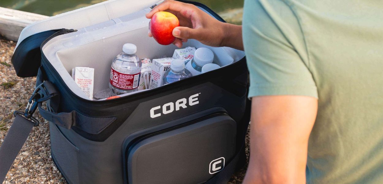 Person sitting by the water with a CORE Climakeep 18 can soft cooler filled with drinks and snacks, featuring shoulder strap and front pocket.