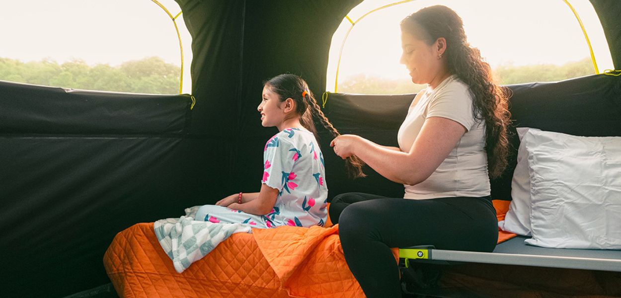 Interior view of a CORE tent with large windows, showing a mother and daughter seated on camping cots with morning light coming in.