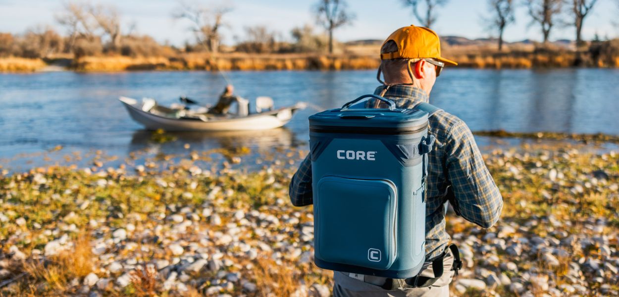 A person with a Navy Climakeep backpack cooler by a lake with another person in a boat.