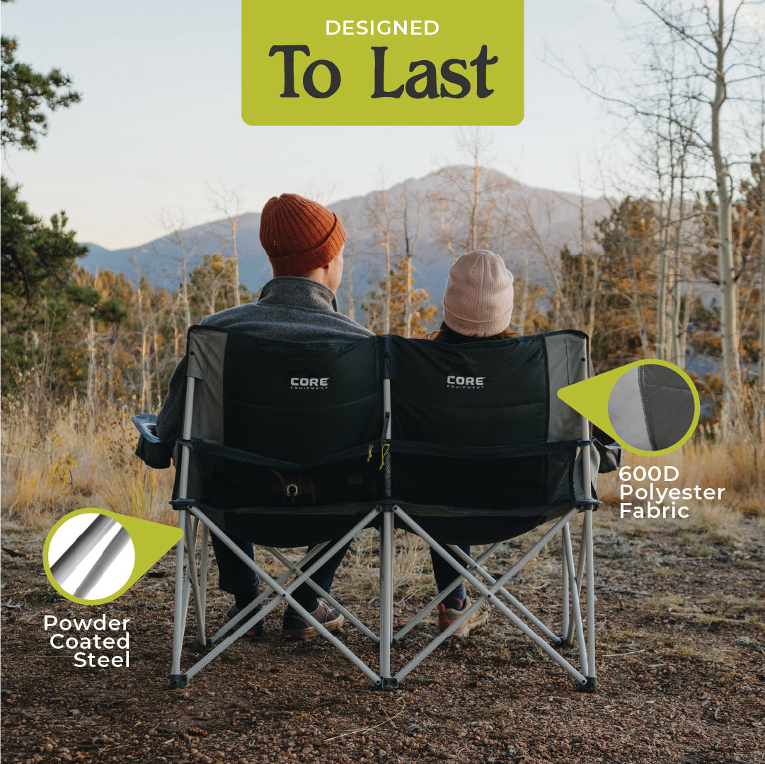 Two people sit side by side in a CORE Loveseat Double Outdoor Camp Chair, facing a scenic view of trees and mountains. Text on the image reads "DESIGNED To Last." Highlights point to features: "Powder Coated Steel" and "600D Polyester Fabric." The brand "CORE" is visible on the chair, perfect for outdoor camping.