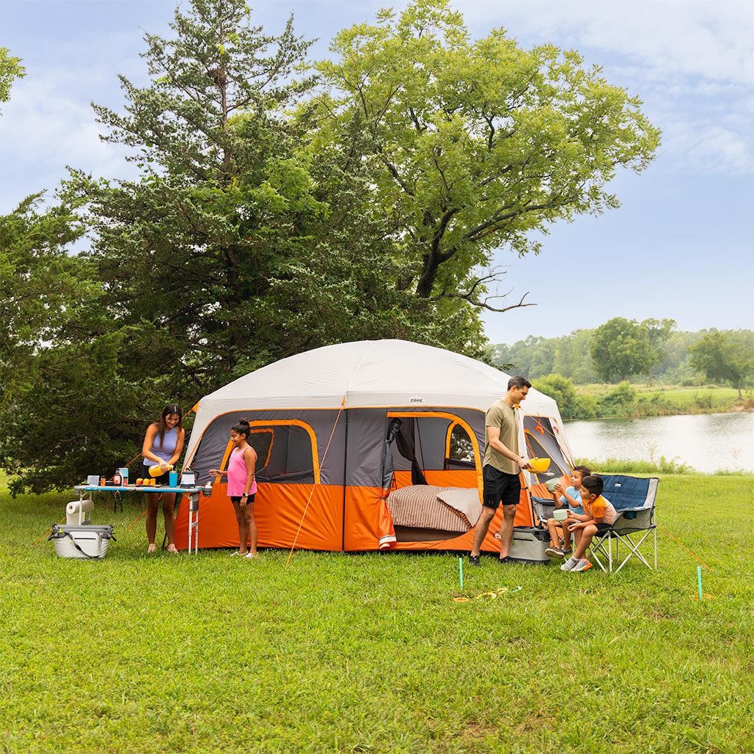 A family is camping outdoors with a CORE 10 Person Straight Wall Cabin Tent in a park setting with a lake in the background.