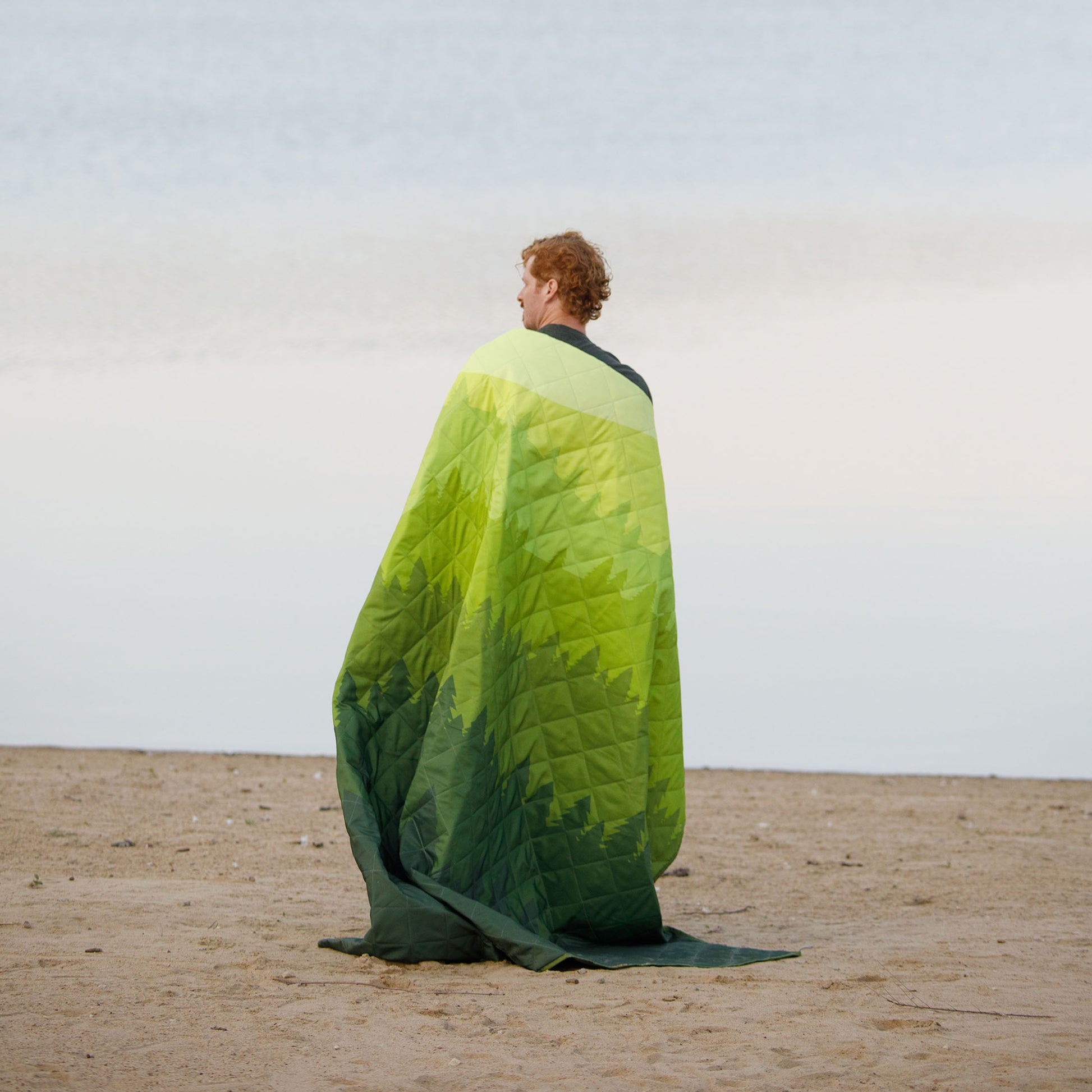 A person with curly hair stands on a sandy beach wrapped in a CORE Staydown Outdoor Travel Weighted Ground Blanket. The beach extends into the background, creating a serene coastal scene.