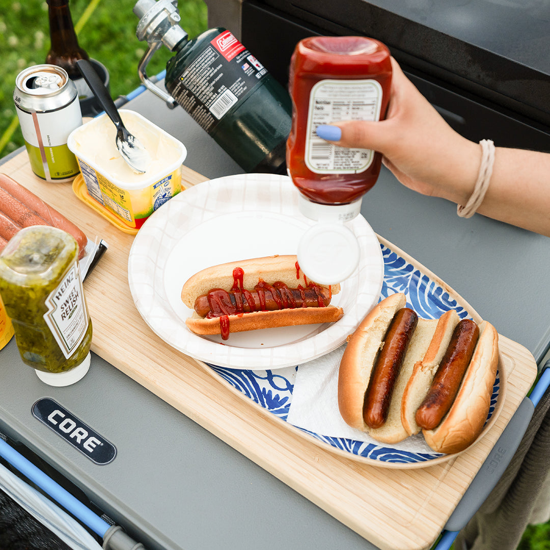 A person is preparing hot dogs with condiments on the CORE Flexrail Camping Table.