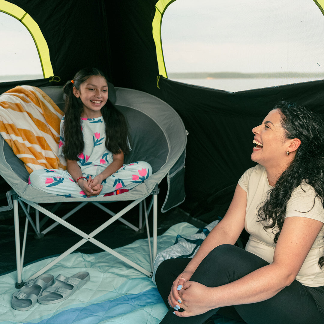 Two people are sitting inside a CORE blockout instant tent, one on a round chair and the other on the ground.