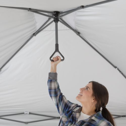 Interior view of a gray canopy tent showing the metal frame structure and center support pole, with a person reaching up to pull a triangular handle used to lock the canopy open, against a plain white background.