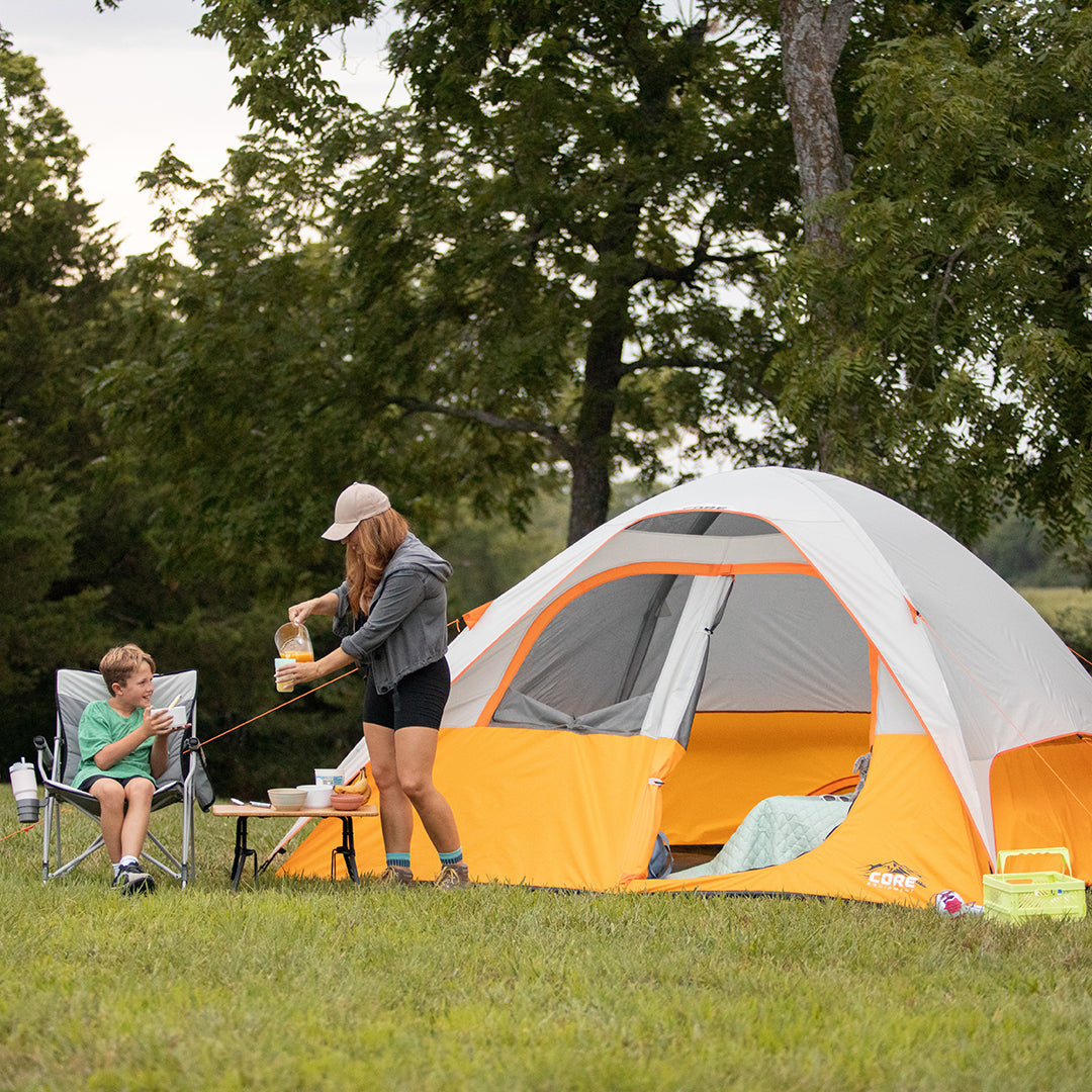 A woman pours a drink at a table beside a CORE orange and gray tent while a boy relaxes in a camping chair, both enjoying an outdoor adventure on grassy ground with trees in the background.