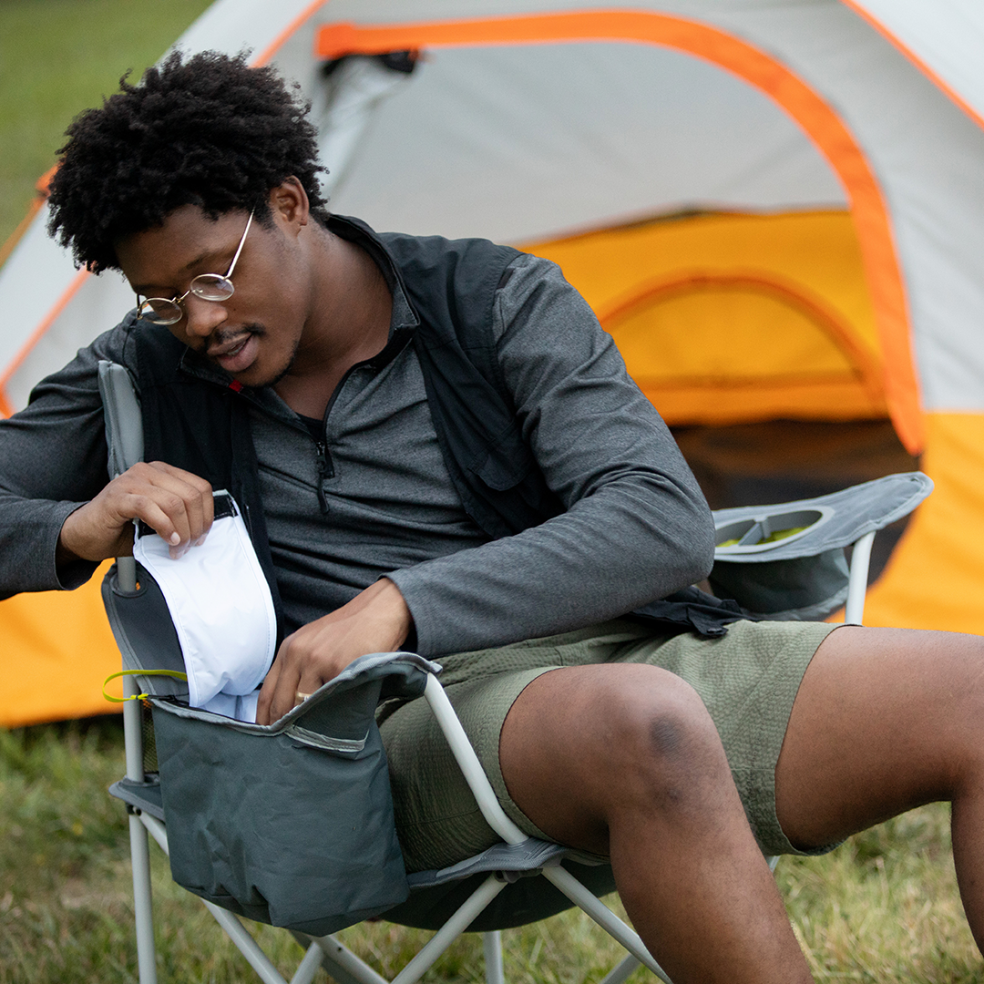 A man enjoys the outdoors on grassy ground, sitting in a CORE cooler quad camping chair outside his tent. He wears glasses, a black jacket, green shorts, and reaches into the chair’s cooler to take out a drink.