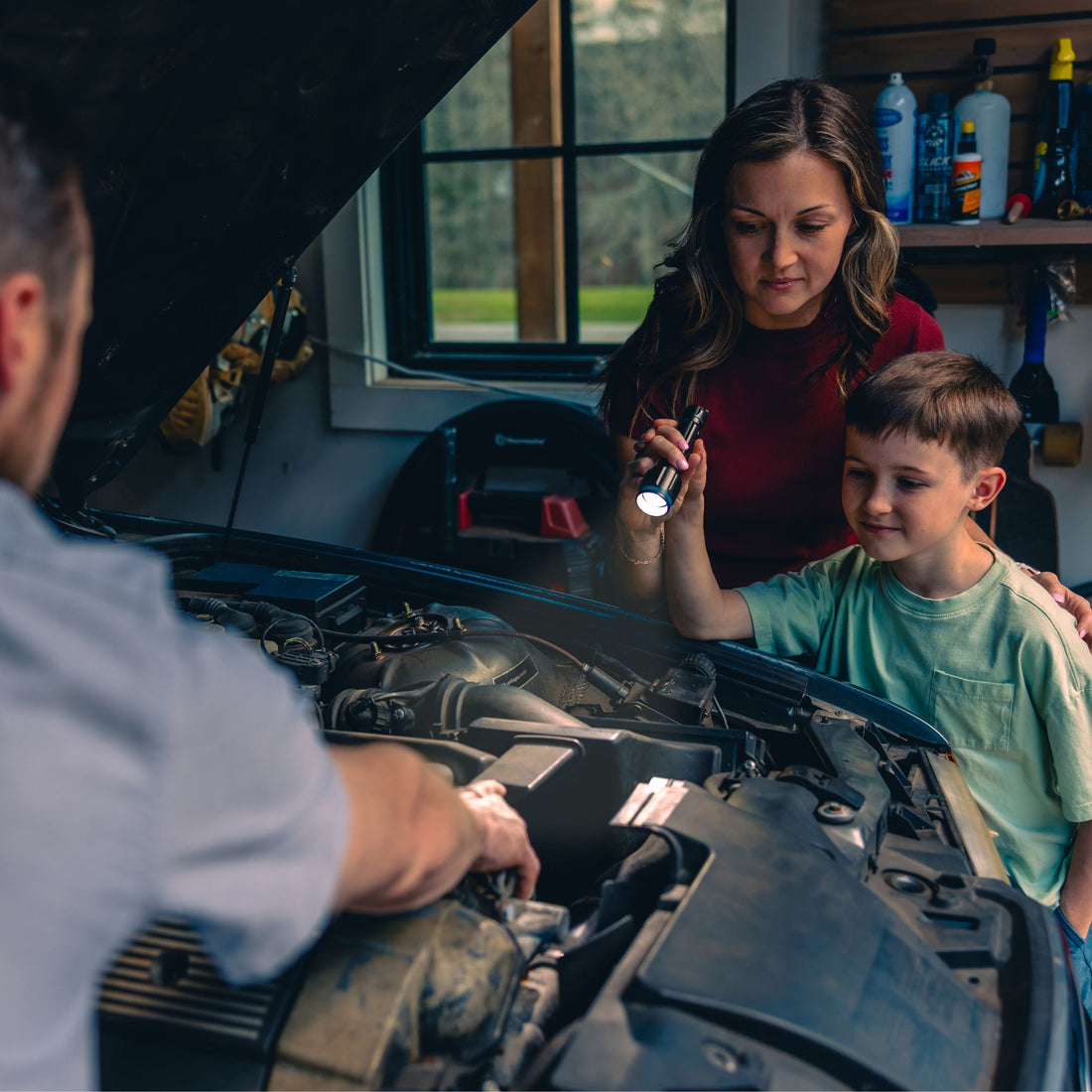 A man works on the engine of a car while a boy points a CORE flashlight and a woman watches. 