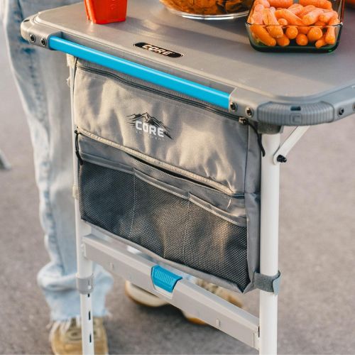 Camping table with CORE FlexRail accessory storage bag attached, holding utensils and gear, with snacks and carrots on a portable outdoor setup