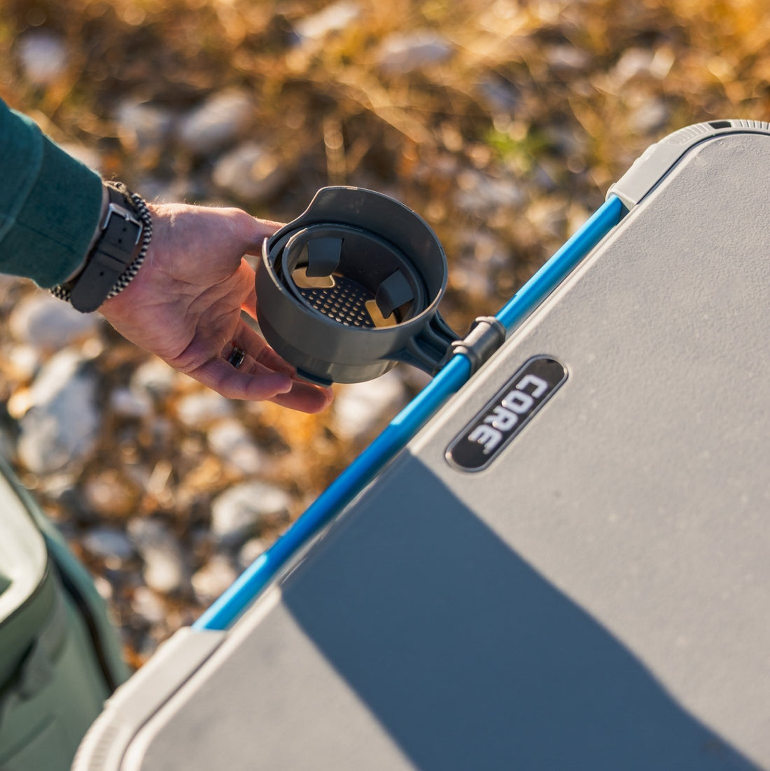 Close-up of hand attaching removable cup holder to CORE FlexRail outdoor table, showing blue accessory rail system along the tabletop edge.