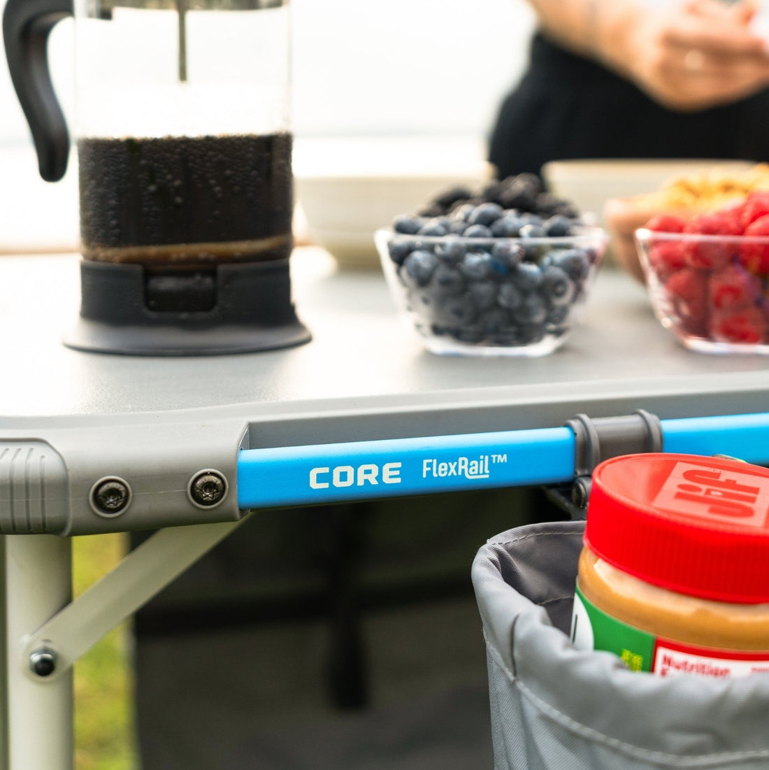 Close-up of CORE FlexRail outdoor table showing blue accessory rail system beneath the tabletop with cup holder and storage bag holding items like food and a coffee press on the surface.
